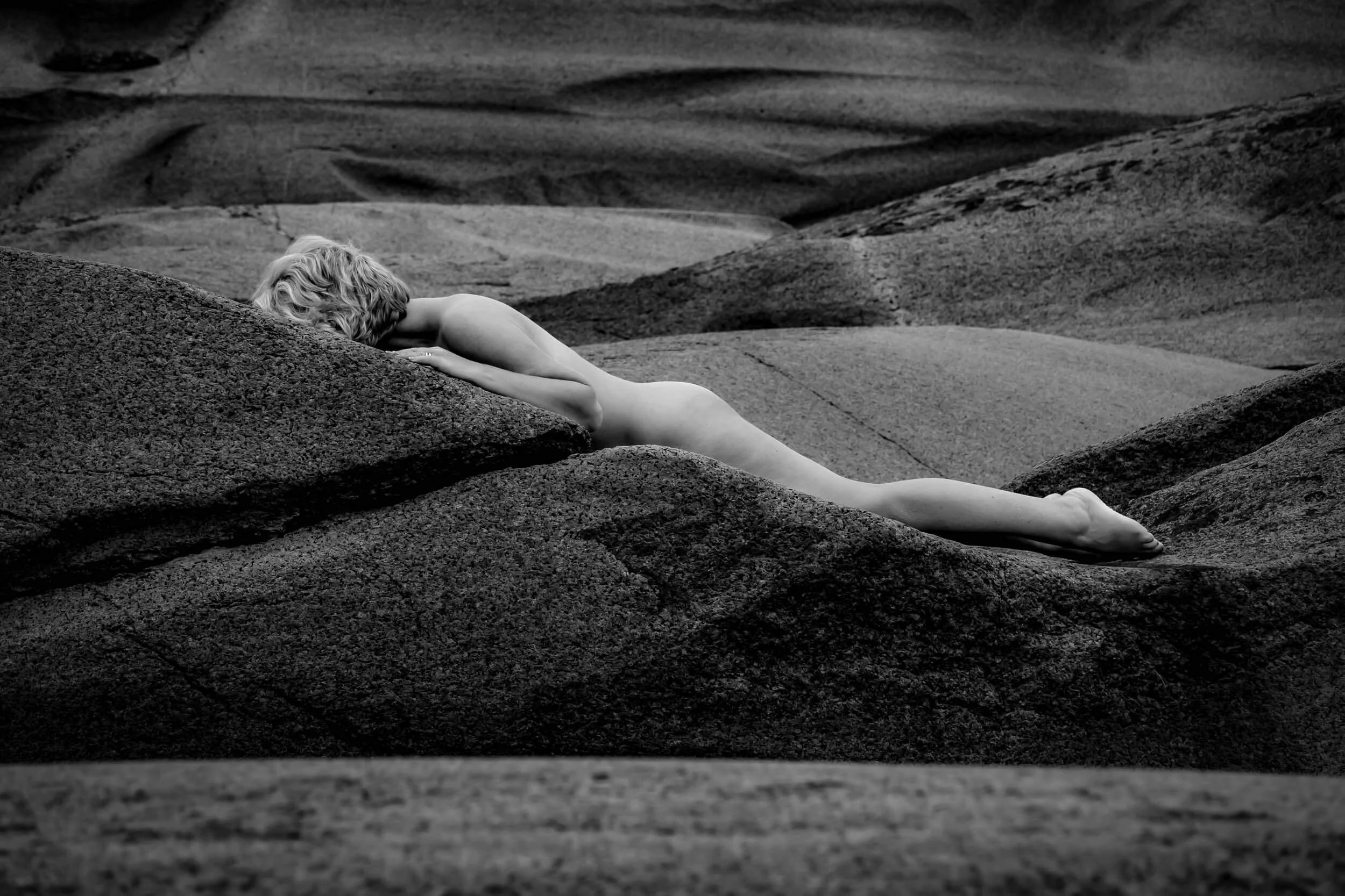 A black and white photograph of a woman lying on rocks with her face resting on her arm. She is surrounded by a rugged, rocky landscape.