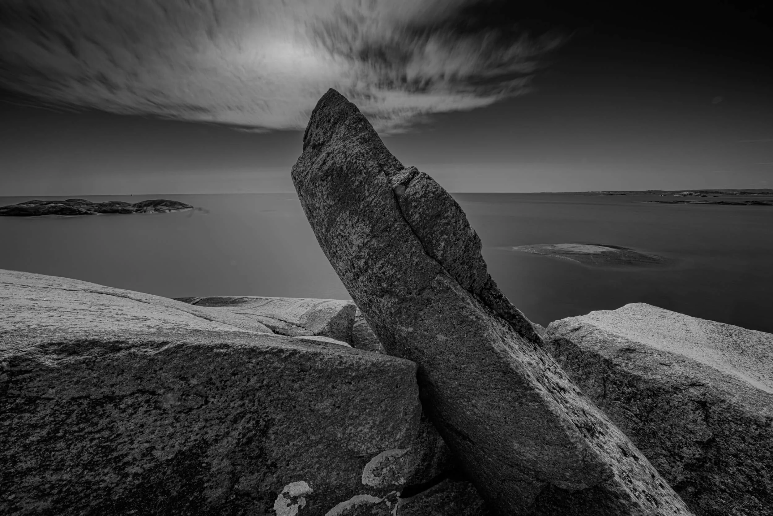 Black and white photo of large rocks and boulders on a shoreline with water and a cloudy sky in the background.
