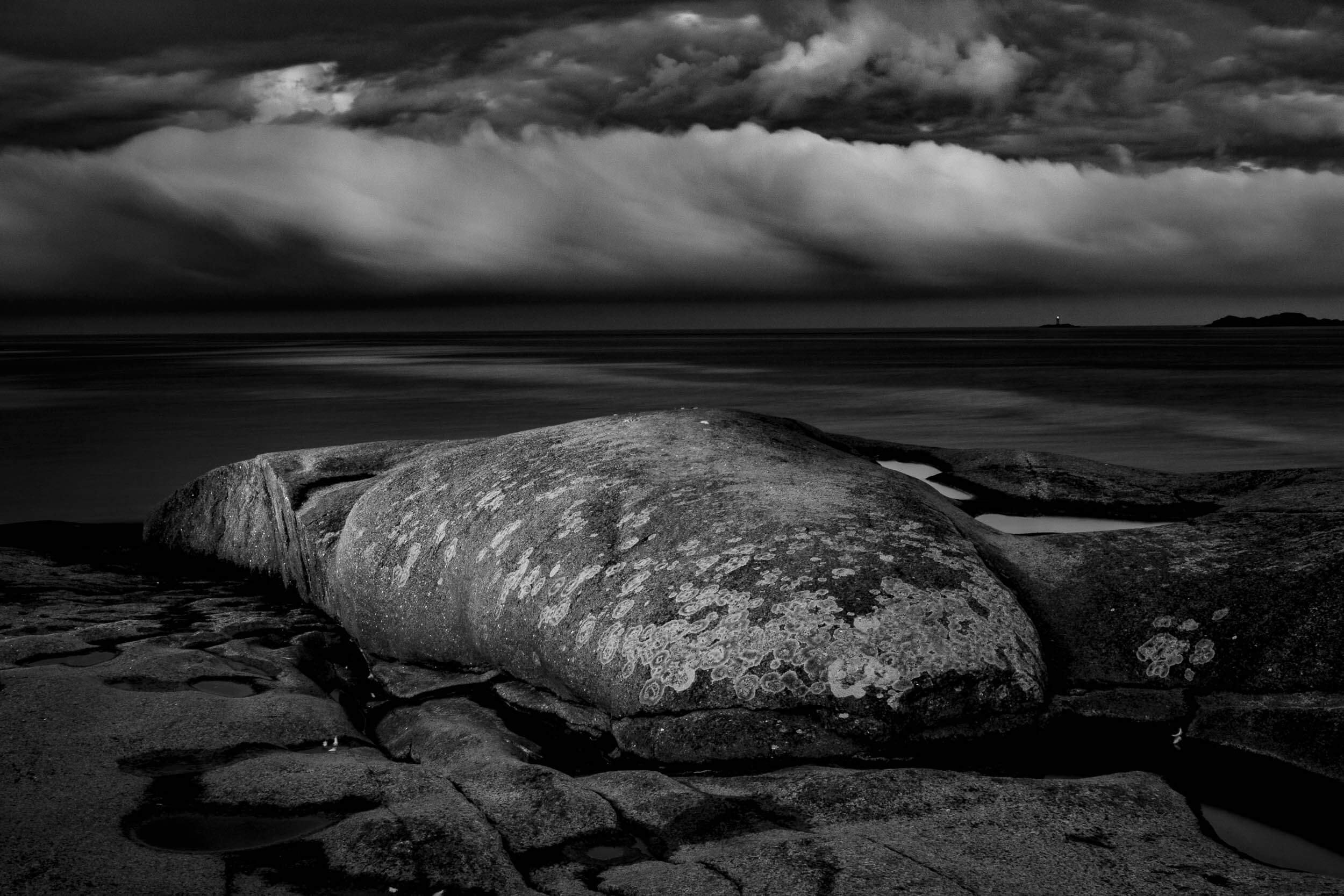 Black and white photo of large rocks and sand on the beach with a cloudy sky and distant lighthouse.
