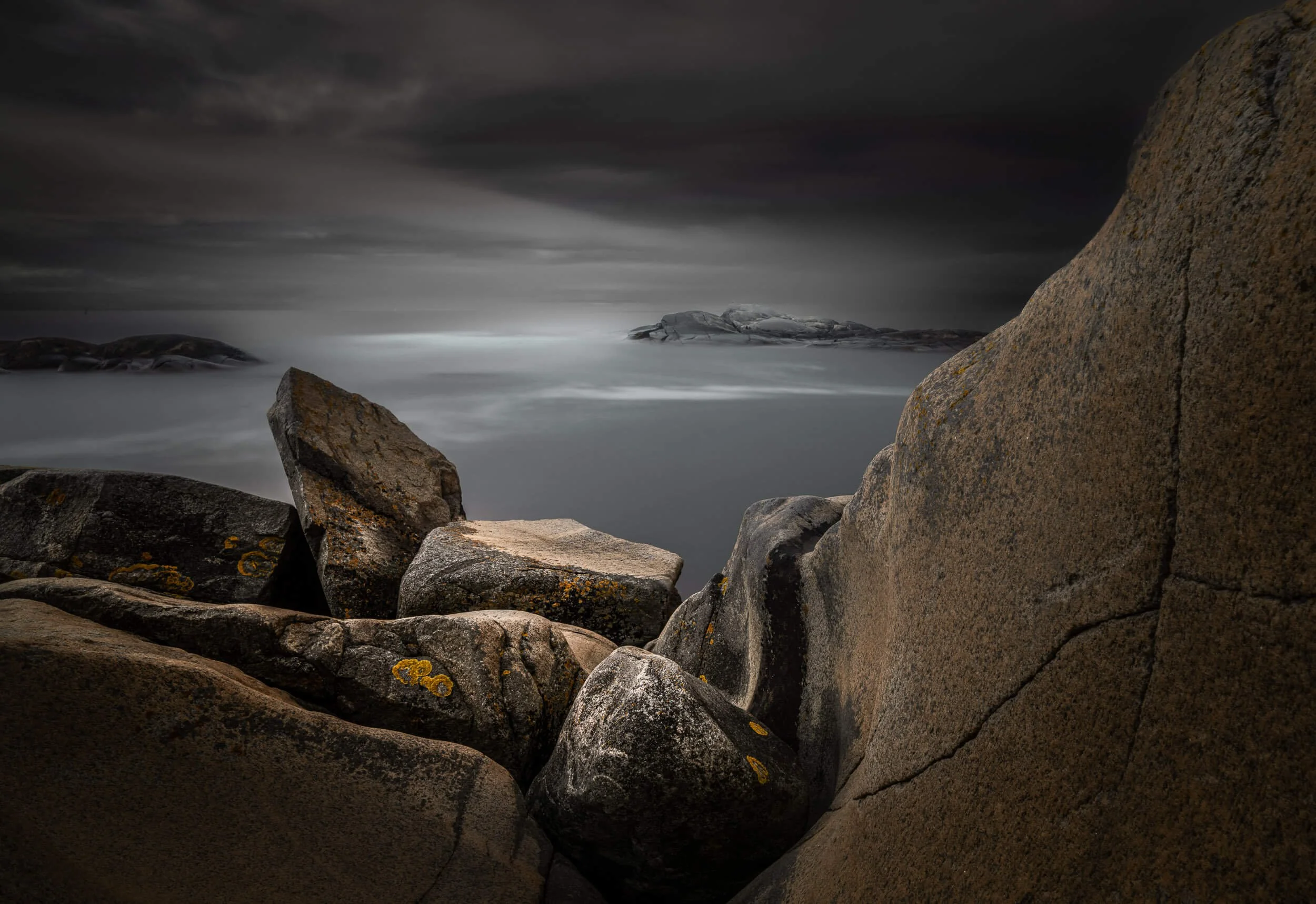 Dark, cloudy sky over rocky shoreline with large boulders and calm water, creating a moody and atmospheric scene.