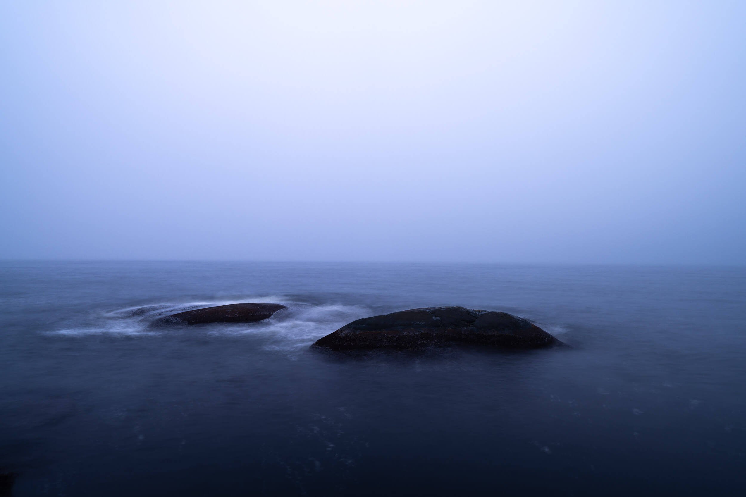 A seascape with large rocks in calm water and a misty sky over the horizon.