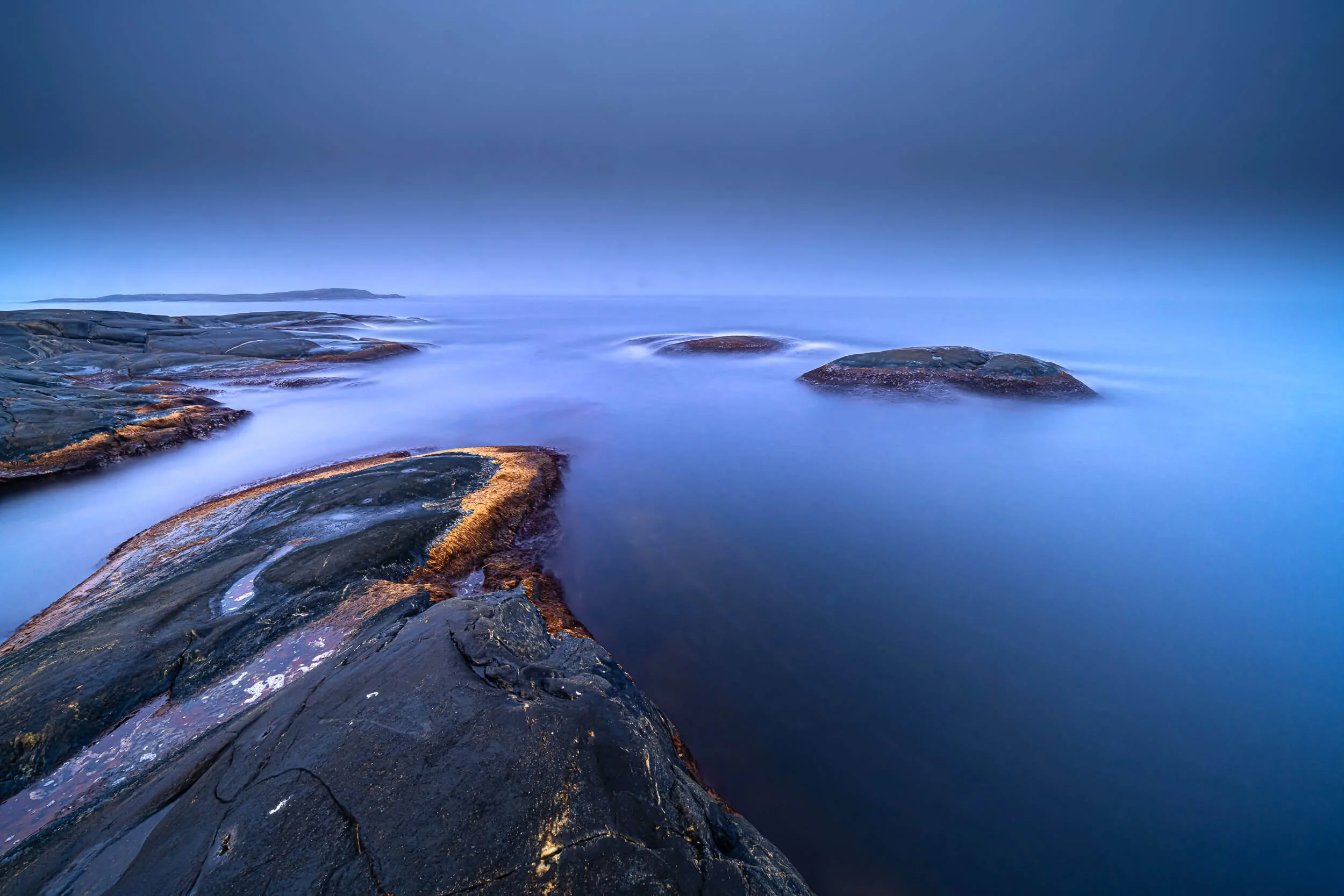 Calm ocean with rocks in the foreground and a distant landmass under a blue sky.