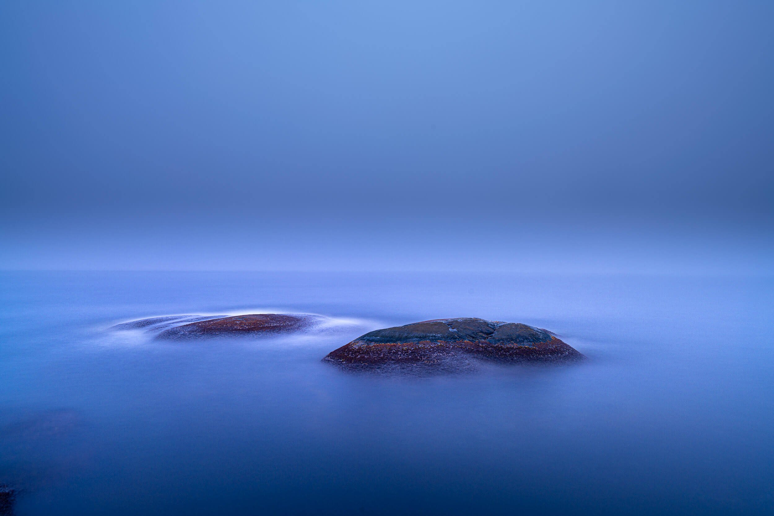 A calm ocean with two large rocks protruding from the water in the foreground. The sky is clear with no clouds, and the overall scene has a tranquil, minimalist aesthetic.