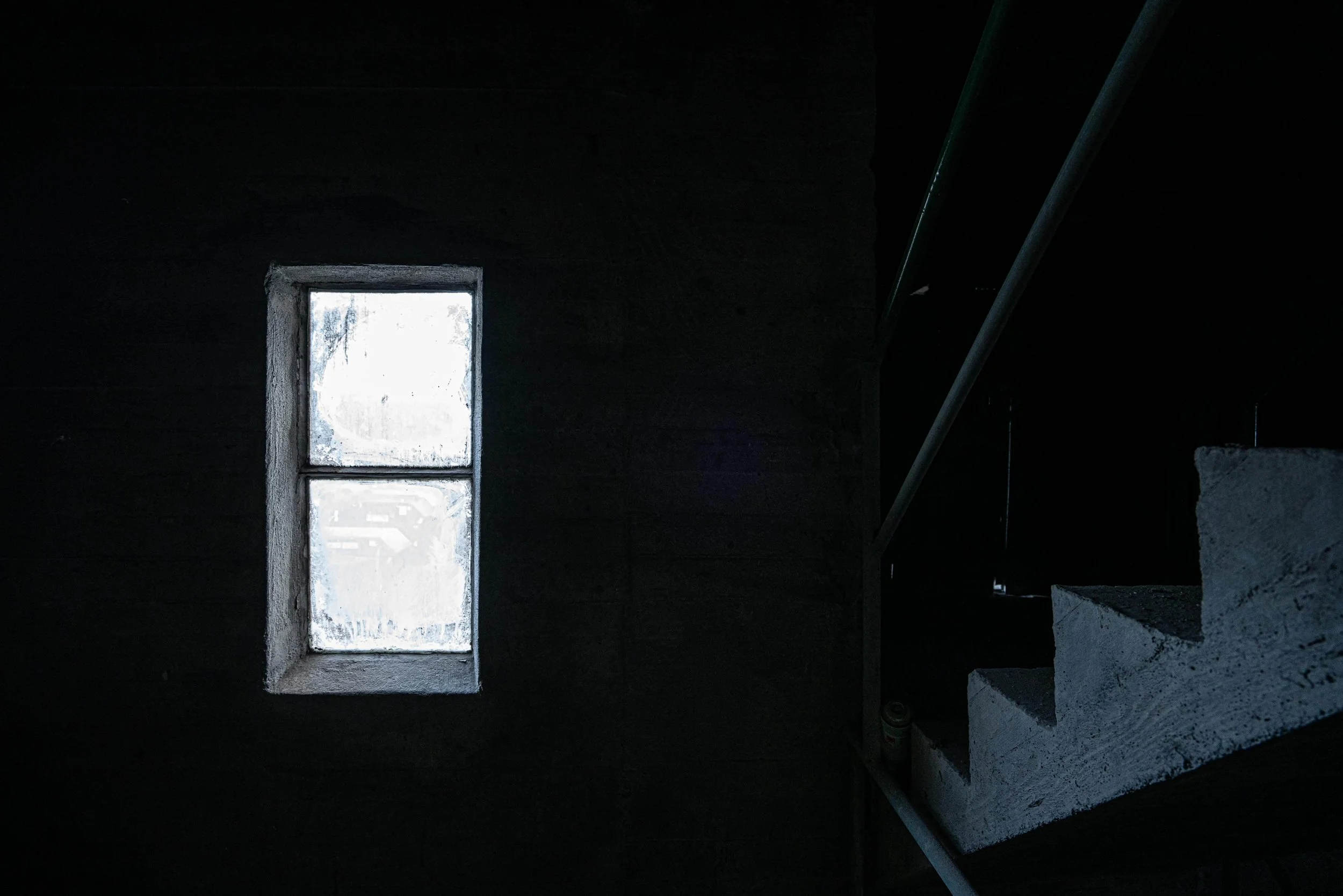 An interior view of a dimly lit room with a window allowing some natural light to enter. The room appears to be unfinished or abandoned, with rough concrete surfaces and stairs leading upward.