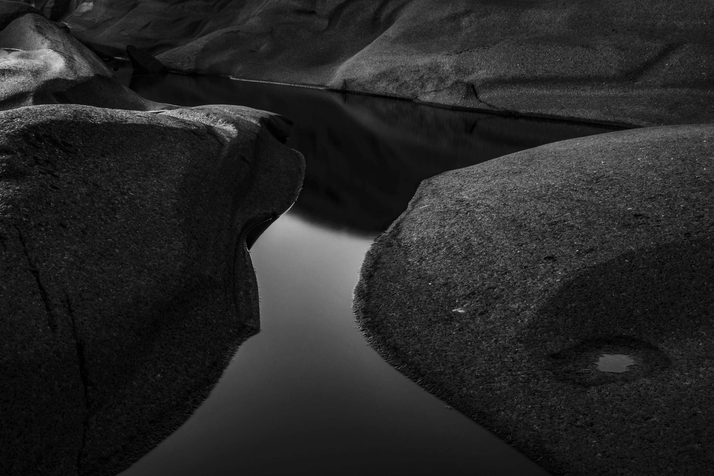 A narrow river or stream flowing between large dark rocks in a black-and-white landscape.