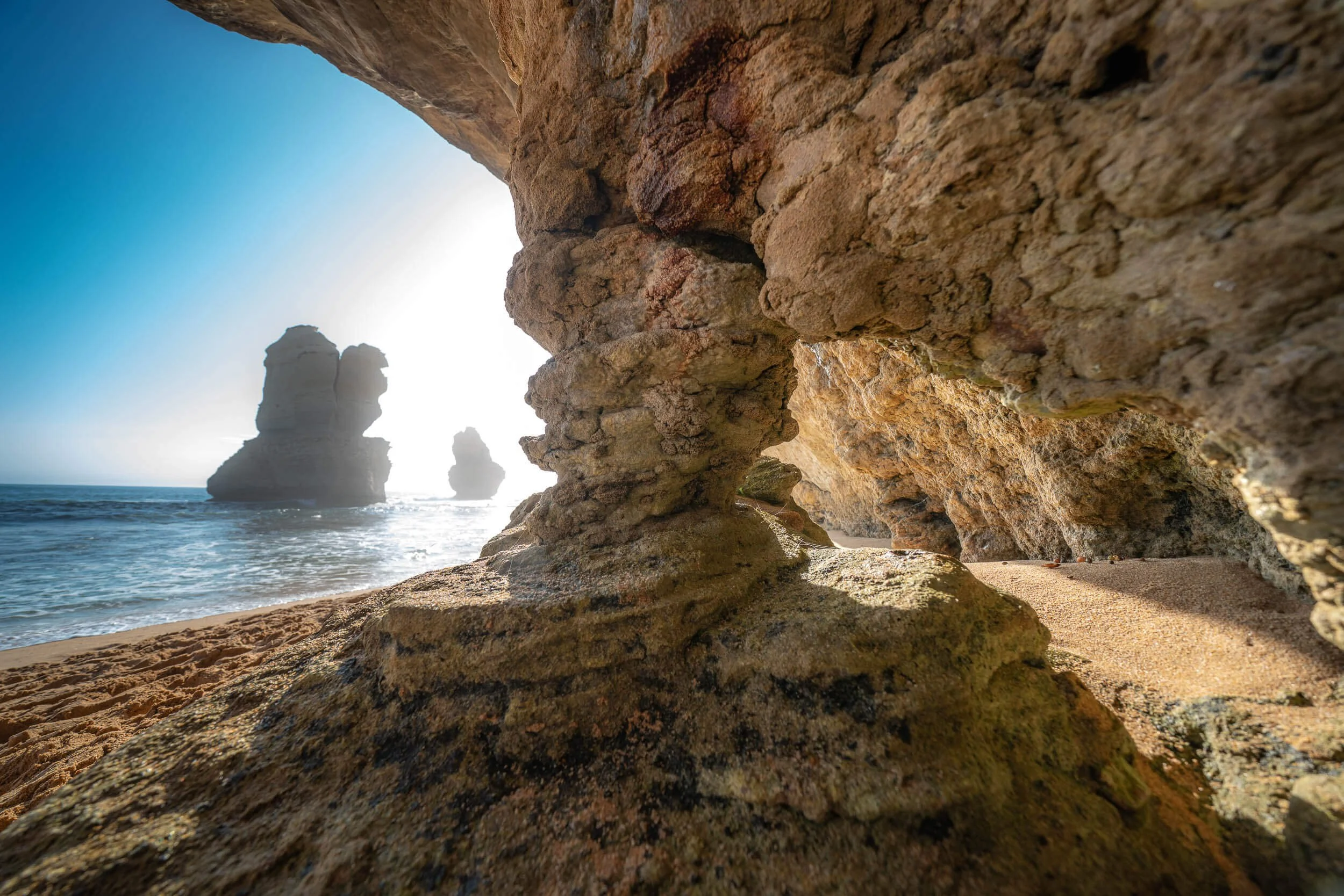 View through a natural rock formation at a beach, showing large rock stacks in the ocean, blue sky, and sunlight.