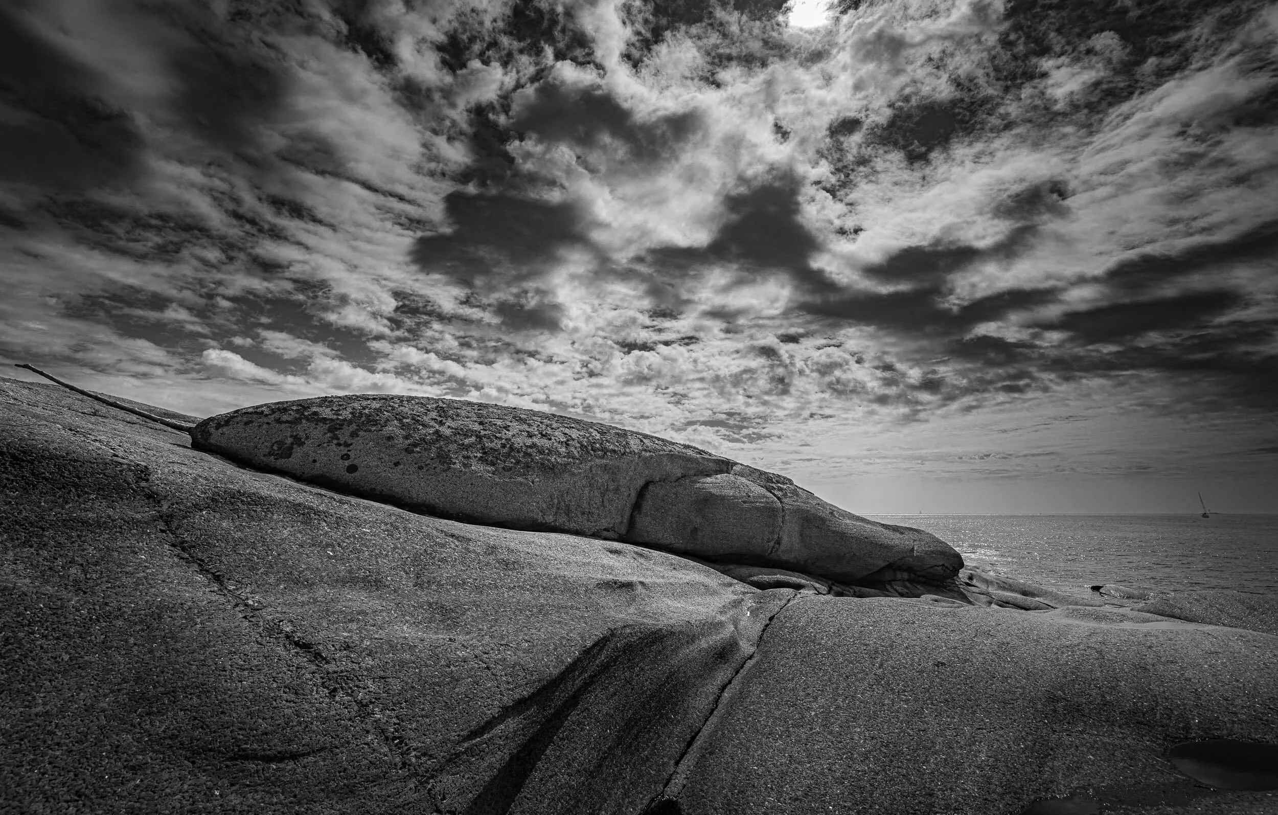 Black and white photograph of a rocky beach with large rounded rocks and clouds in the sky extending to the horizon.