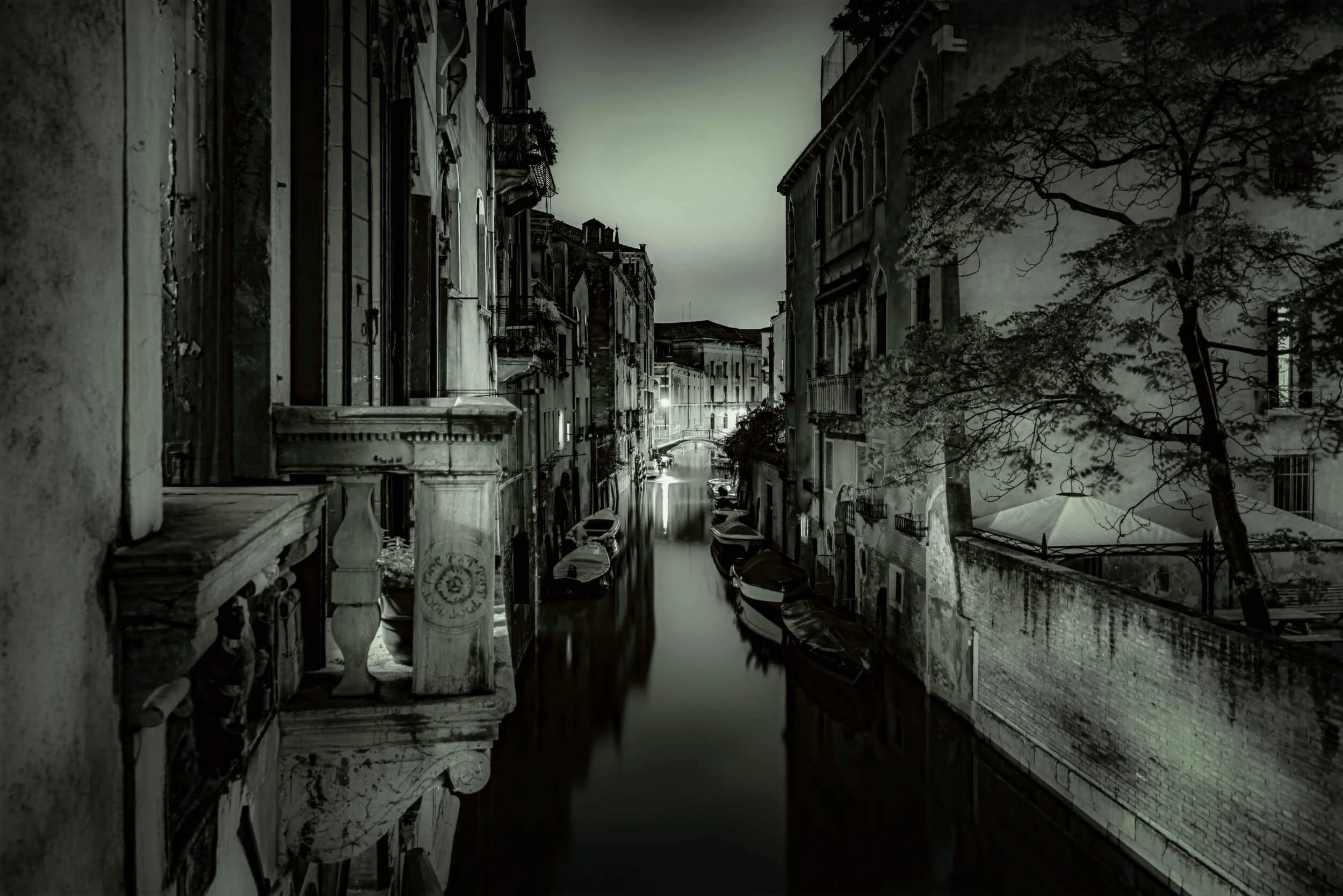 A black and white photograph of a narrow canal in Venice, Italy, with boats docked along the sides and old buildings with balconies lining both sides of the water, under a cloudy sky.