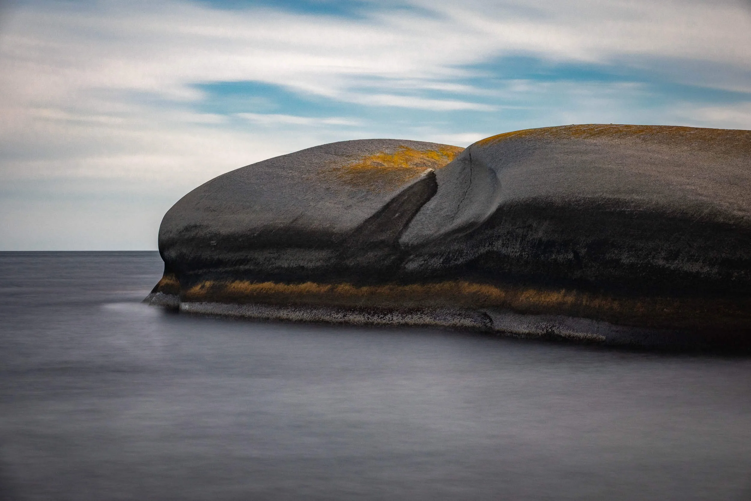Large smooth rock formation along a shoreline with a cloudy sky overhead.