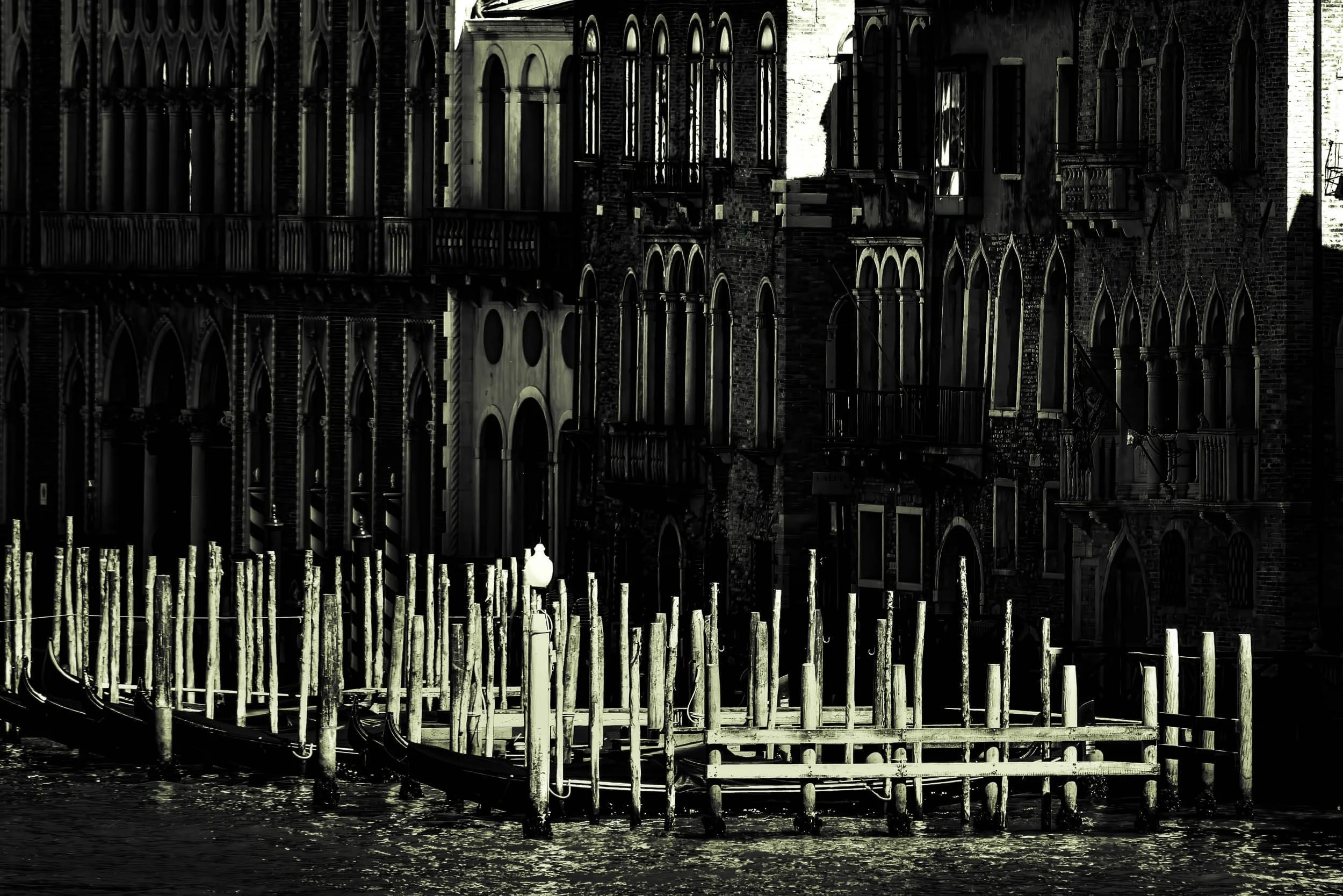 Black and white photo of buildings with arched windows and a series of wooden posts and docks along a waterway.