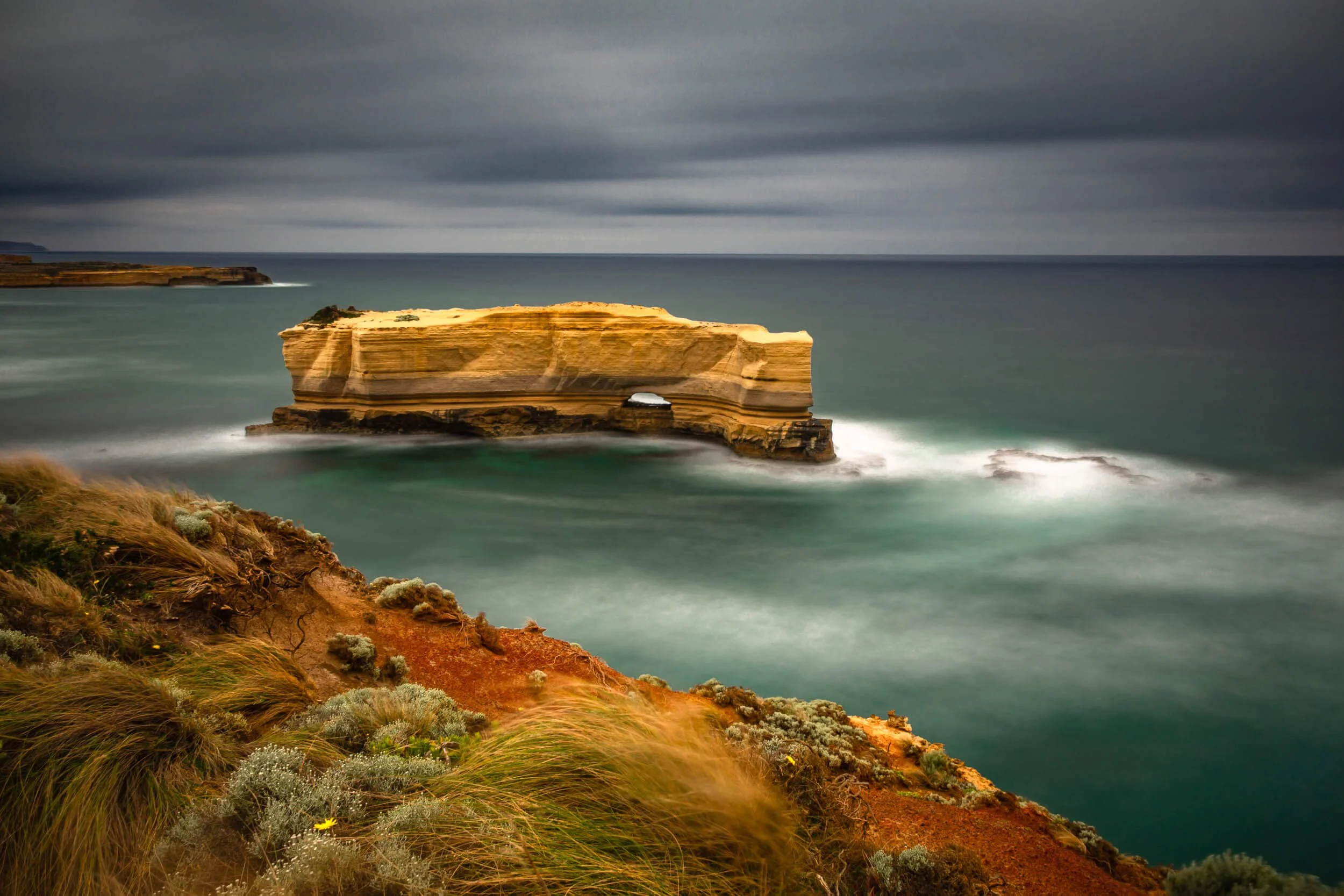 A scenic coastal landscape with a large, layered rock formation surrounded by water, grassy coastal vegetation, and distant cliffs under a cloudy sky.
