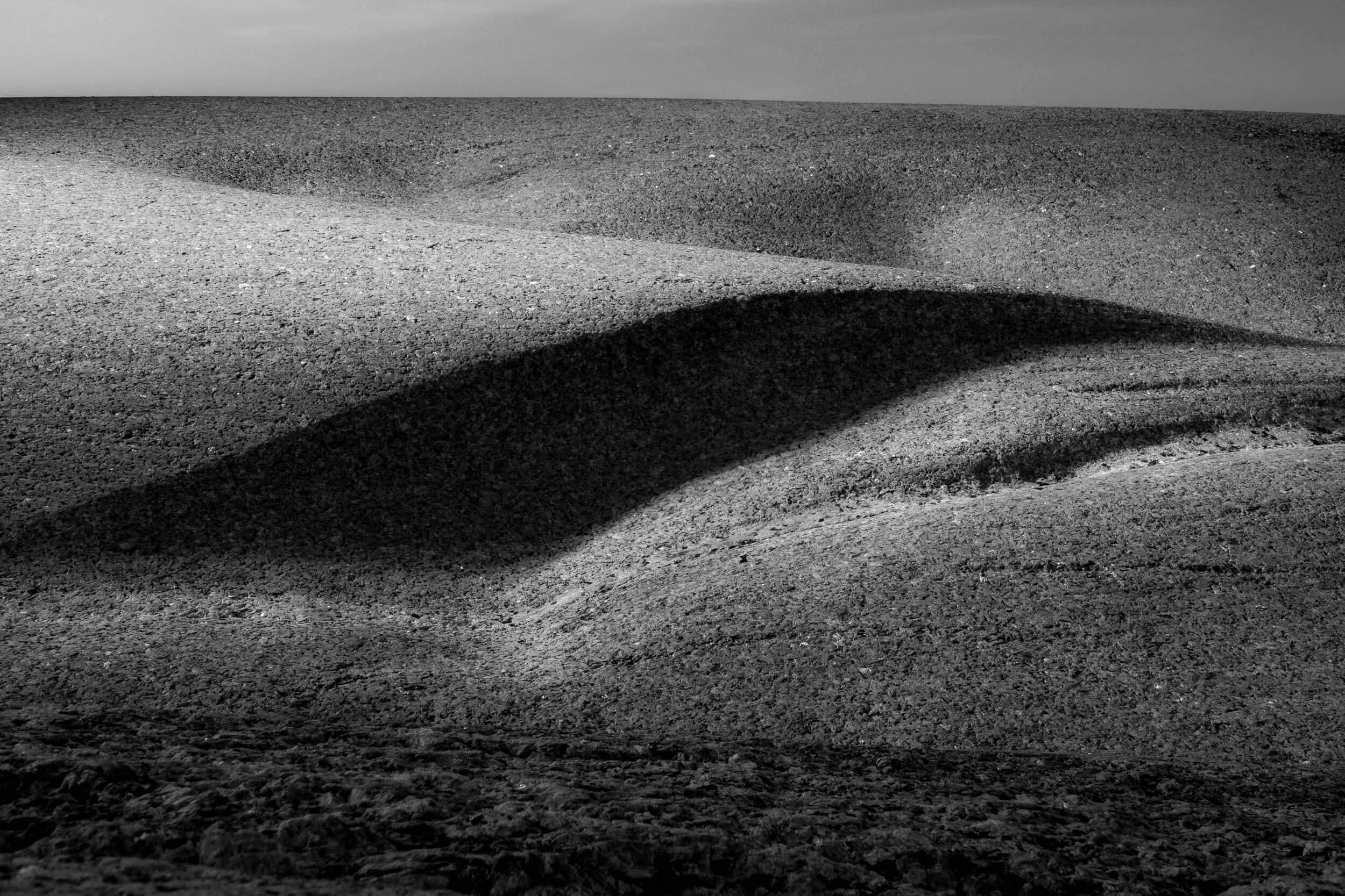 A close-up black and white photograph of rippled and textured sand dunes or landscape with shadows and a cloudy sky in the background.
