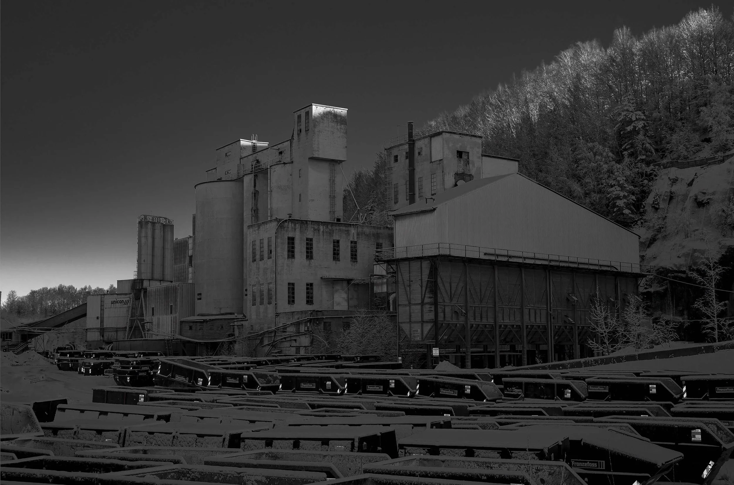 Black and white photo of an old industrial building with multiple sections, surrounded by trees, and train tracks in the foreground.