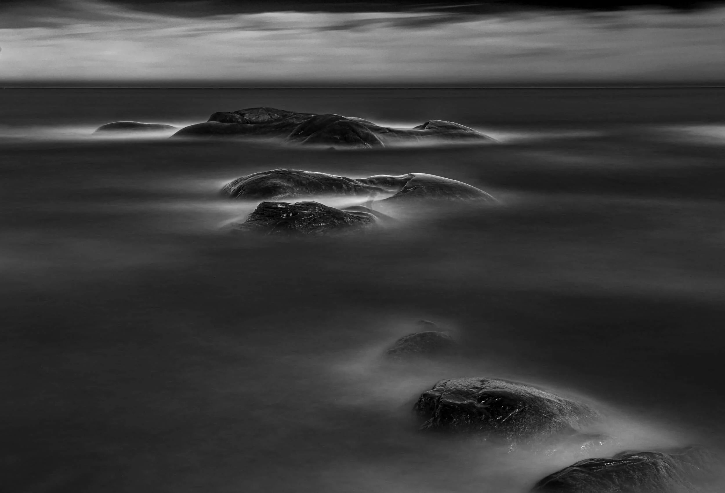 Black and white long exposure photograph of rocks in a calm body of water with a cloudy sky in the background.