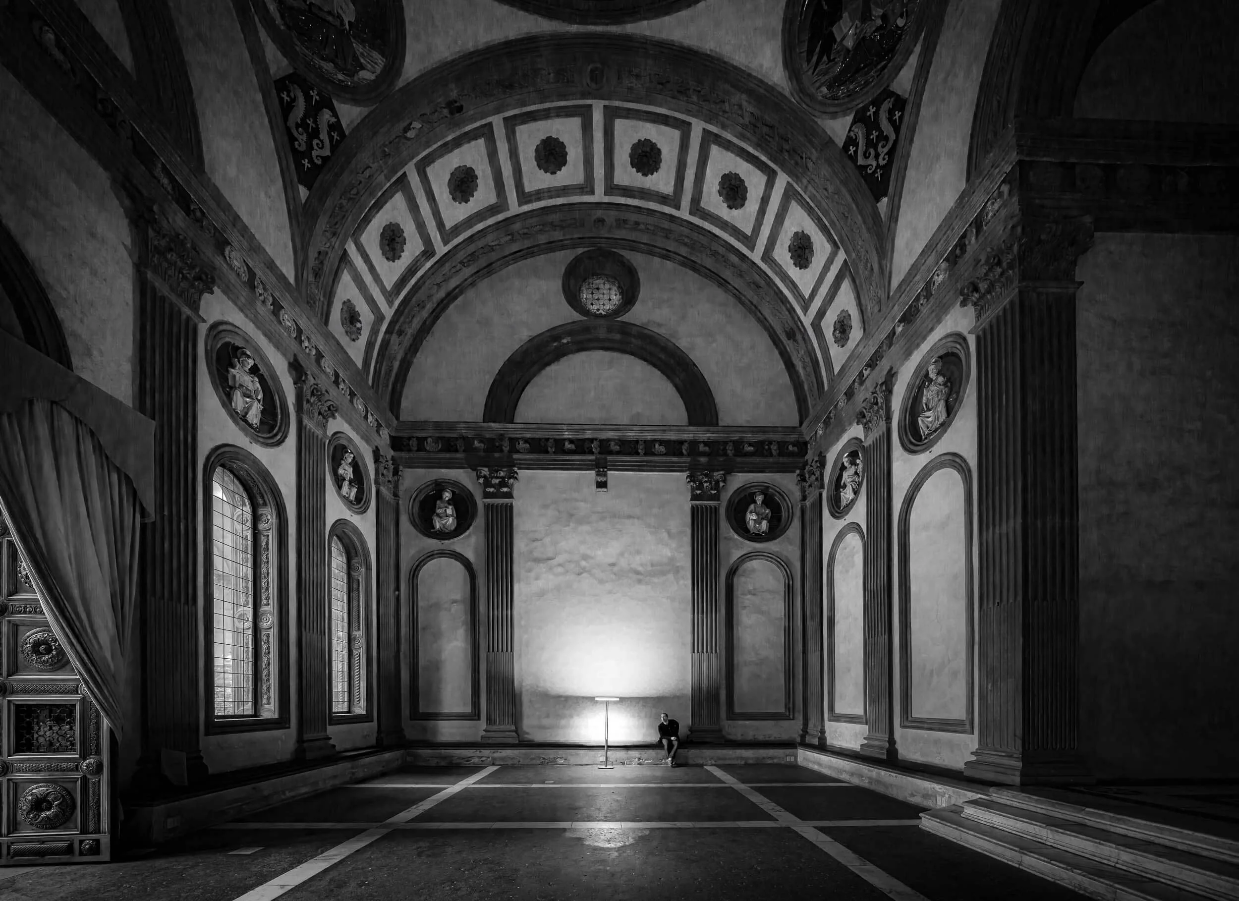 A large, ornate hall with high, vaulted ceilings and decorative arches. There are tall, narrow windows on the left, and intricate architectural details on the walls and columns. A person is sitting on the floor near a lamp in the distance.