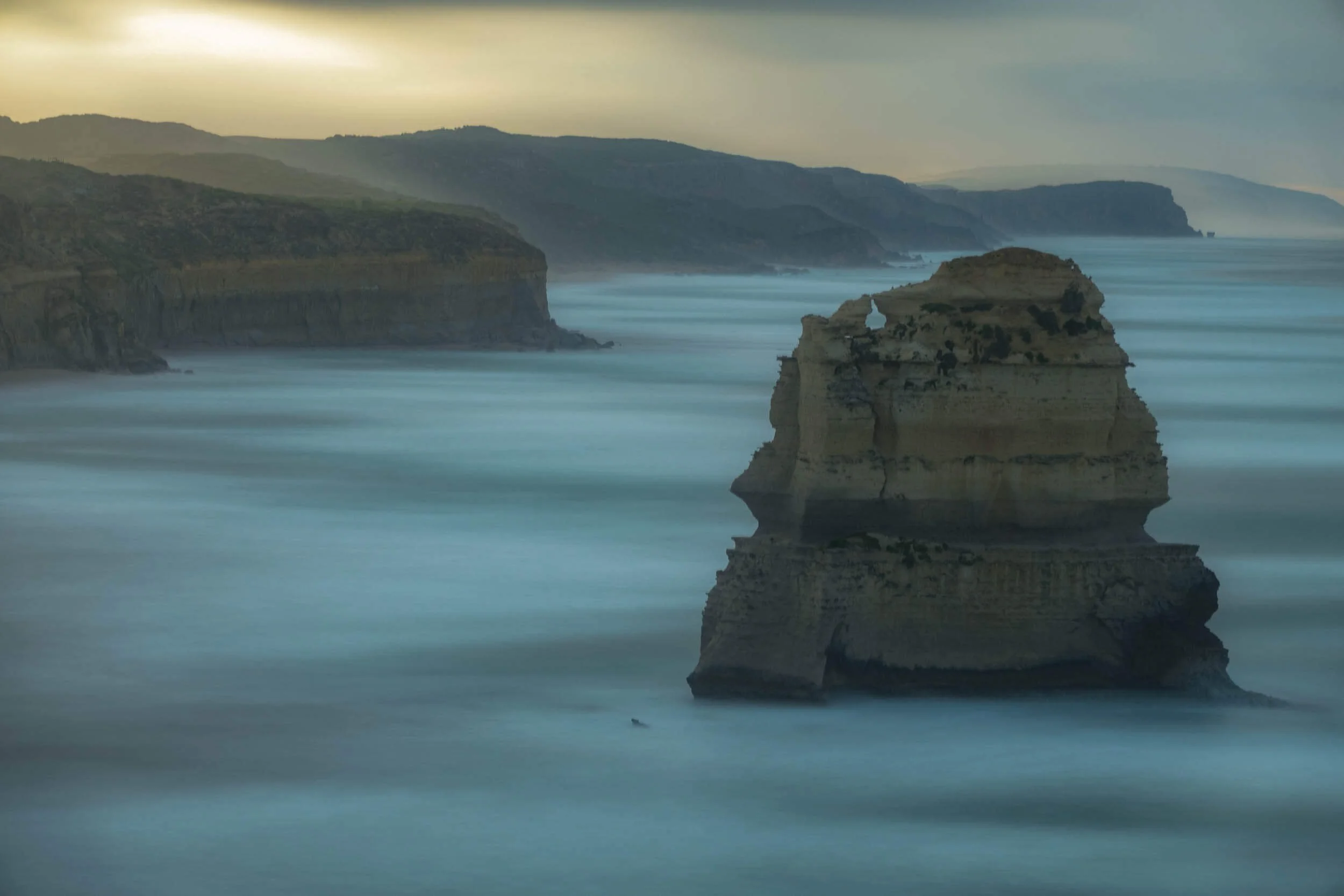 Cliffs and rock formations along a rugged coastline, with the ocean in the foreground and distant hills under a cloudy sky.