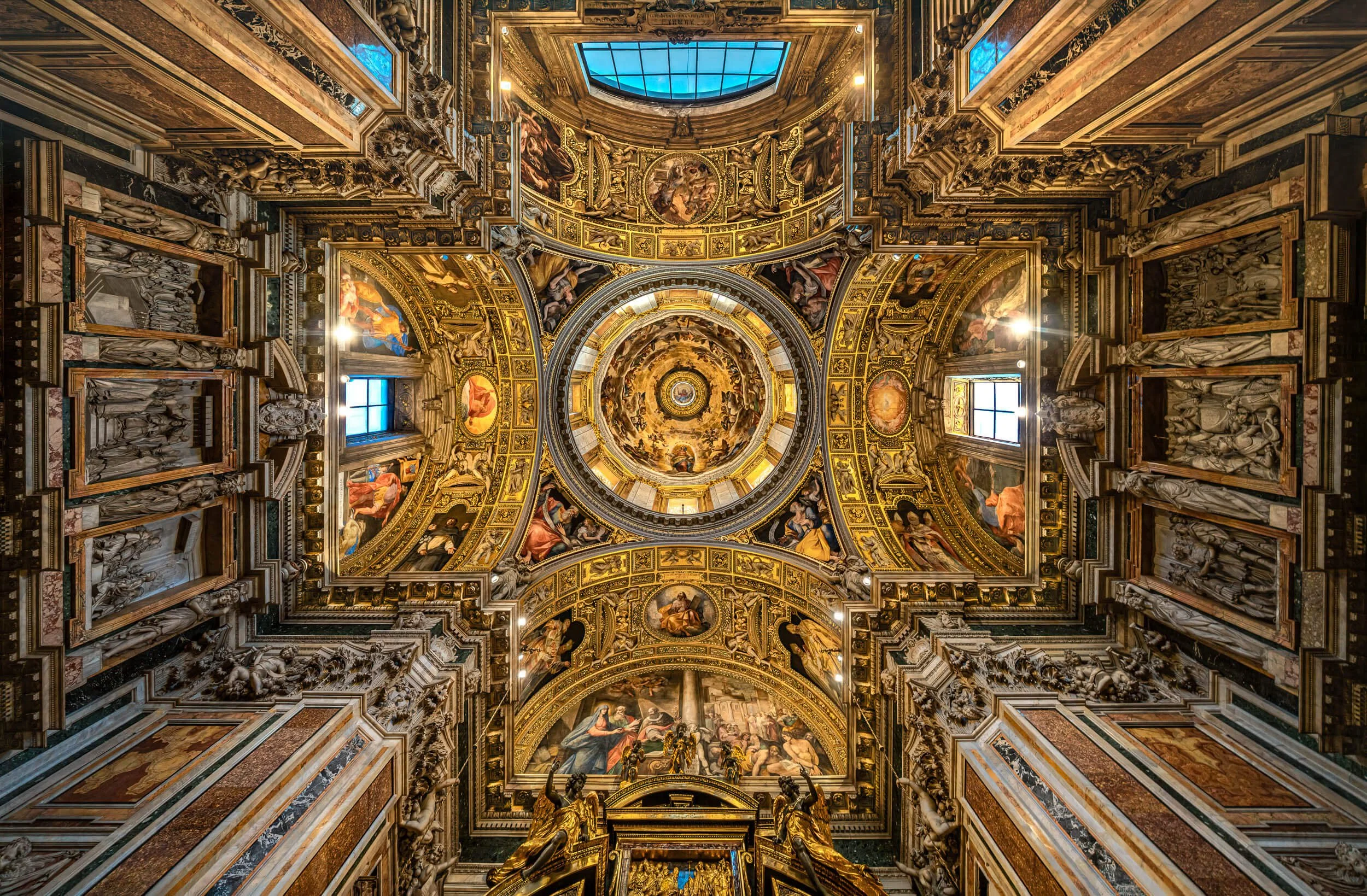 The ornate interior of a grand cathedral or basilica, featuring a richly decorated ceiling with religious artwork, sculptures, and gold accents, as viewed from the ground looking up.