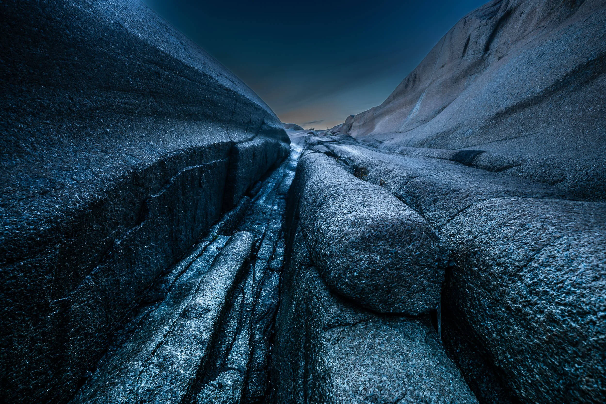 A narrow rocky canyon with smooth, dark gray and blue rocks, under a deep blue sky at dusk or dawn.