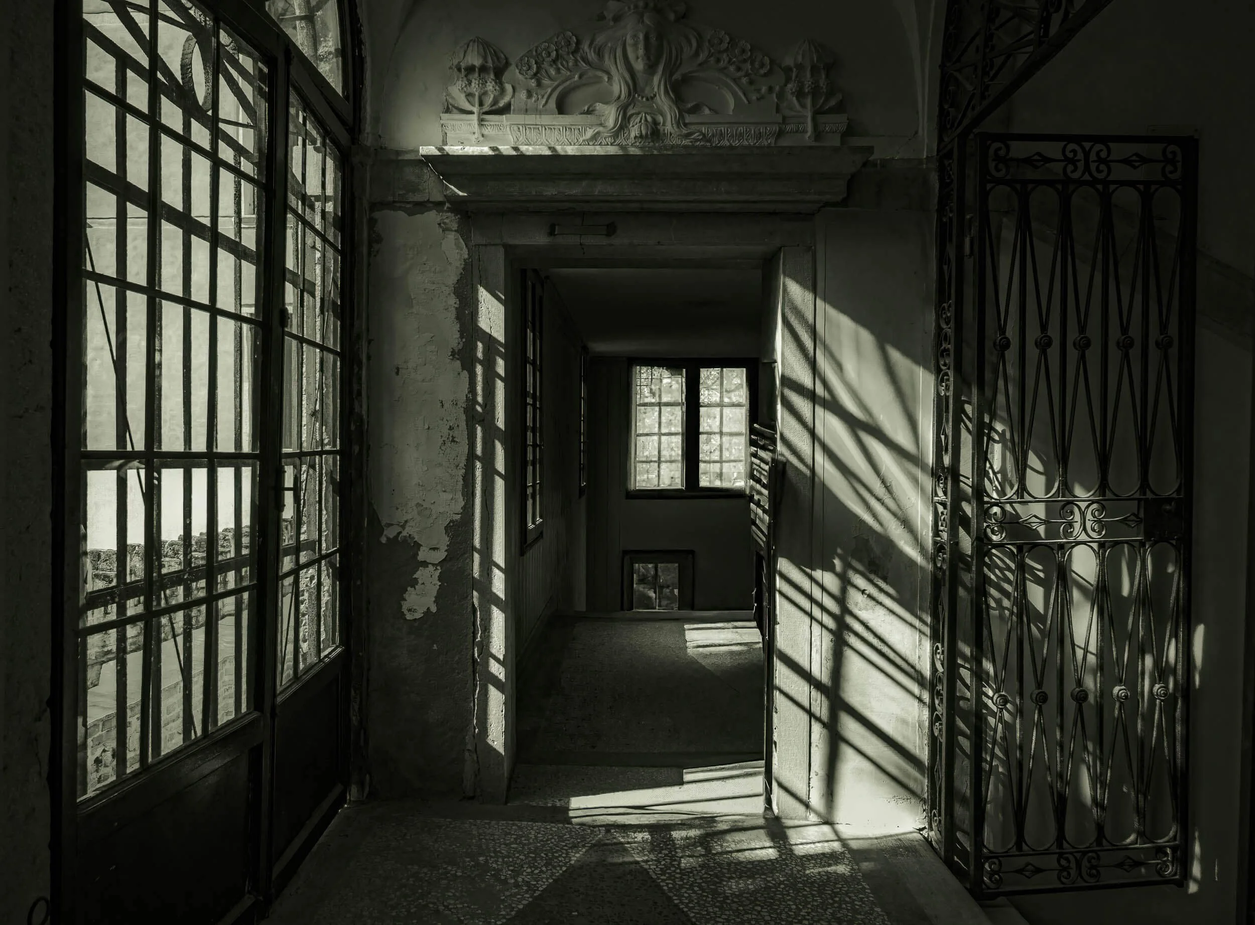 An old, abandoned hallway with barred iron gates, peeling plaster walls, dusty windows casting shadows, and a decorative relief sculpture at the top of the wall.