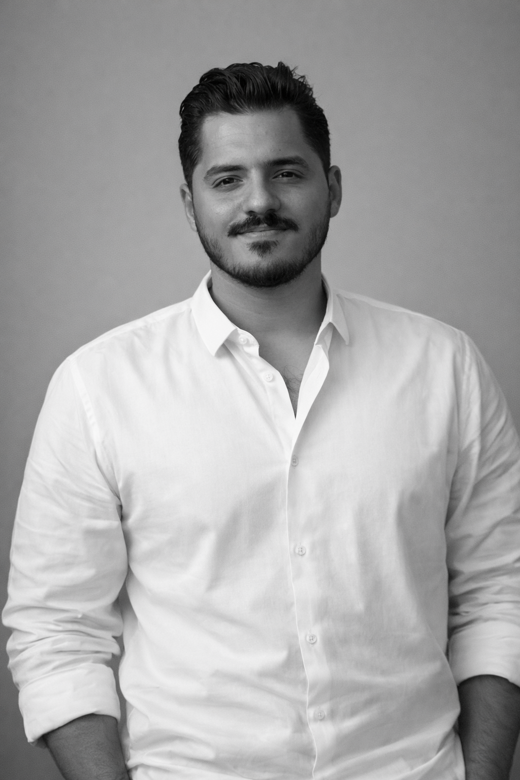 Black and white portrait of a man wearing a white shirt with the top button undone, standing against a plain background with a slight smile.