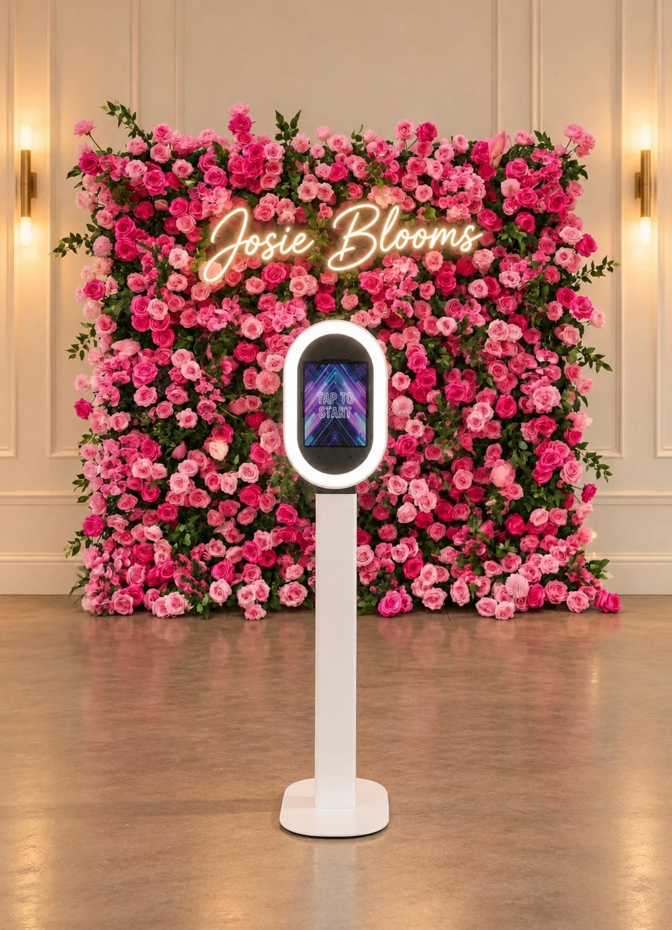 A floral display with pink roses and greenery behind a stand with a screen that reads 'Tap to Start' and a neon sign that says 'Josie Blooms.'