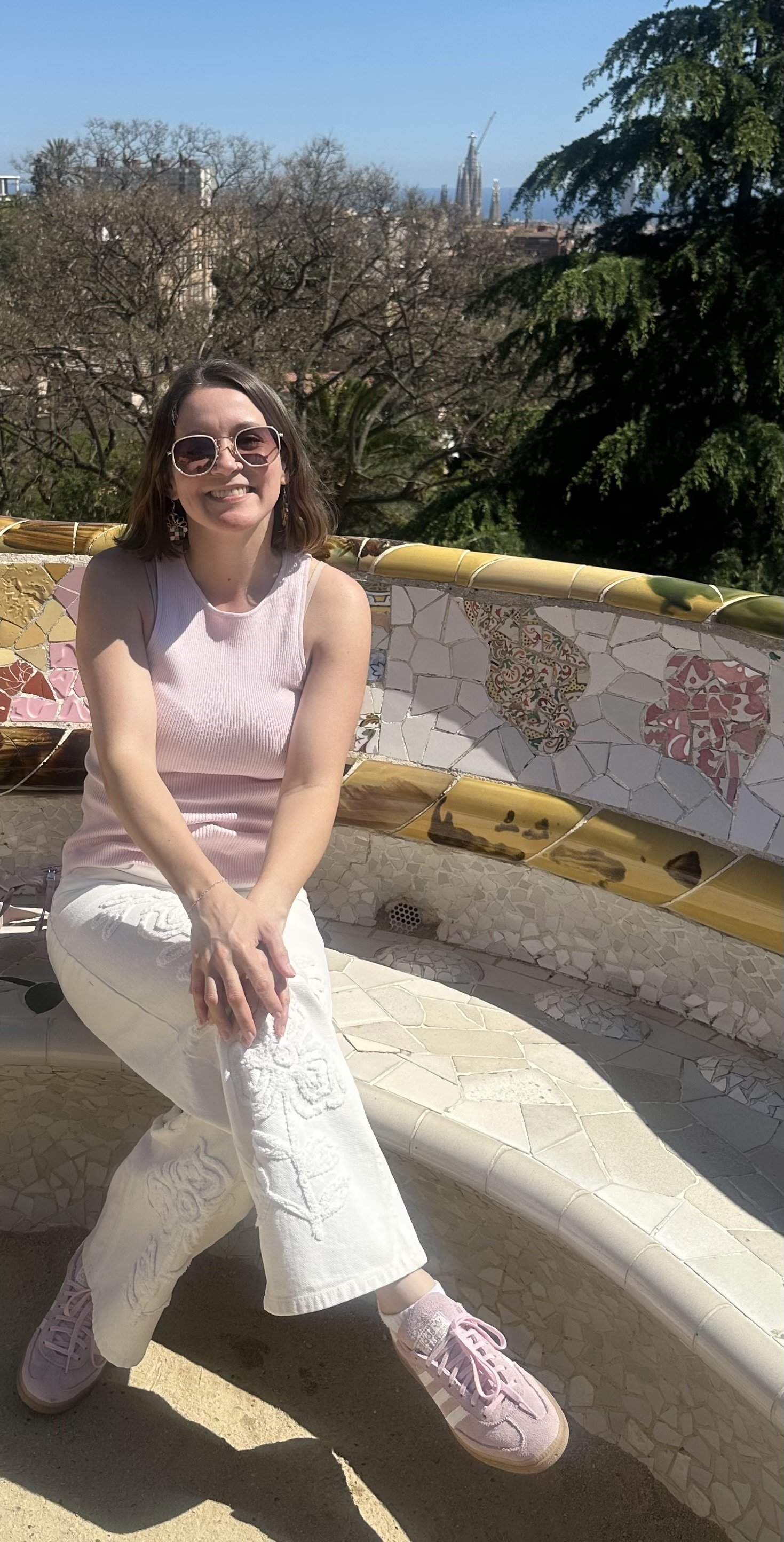 A woman wearing sunglasses smiling and sitting on a tiled bench outdoors in front of trees with a cityscape and skyscraper in the distance.