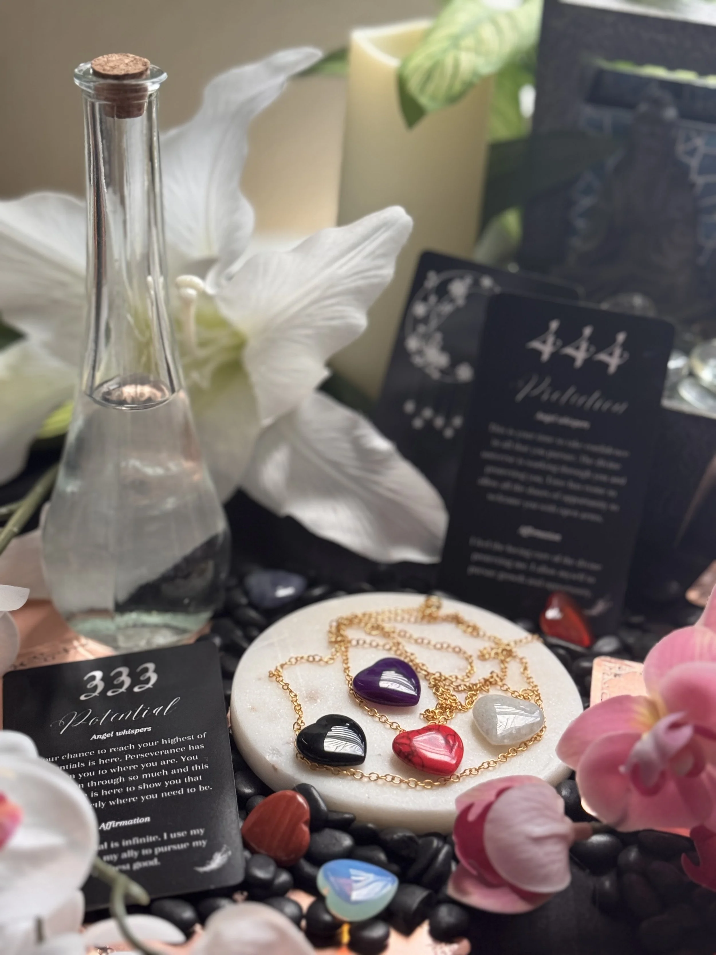 Jewelry display with colorful glass pendants, a gold chain necklace, and a purple heart-shaped pendant, surrounded by pink flowers and small black stones.