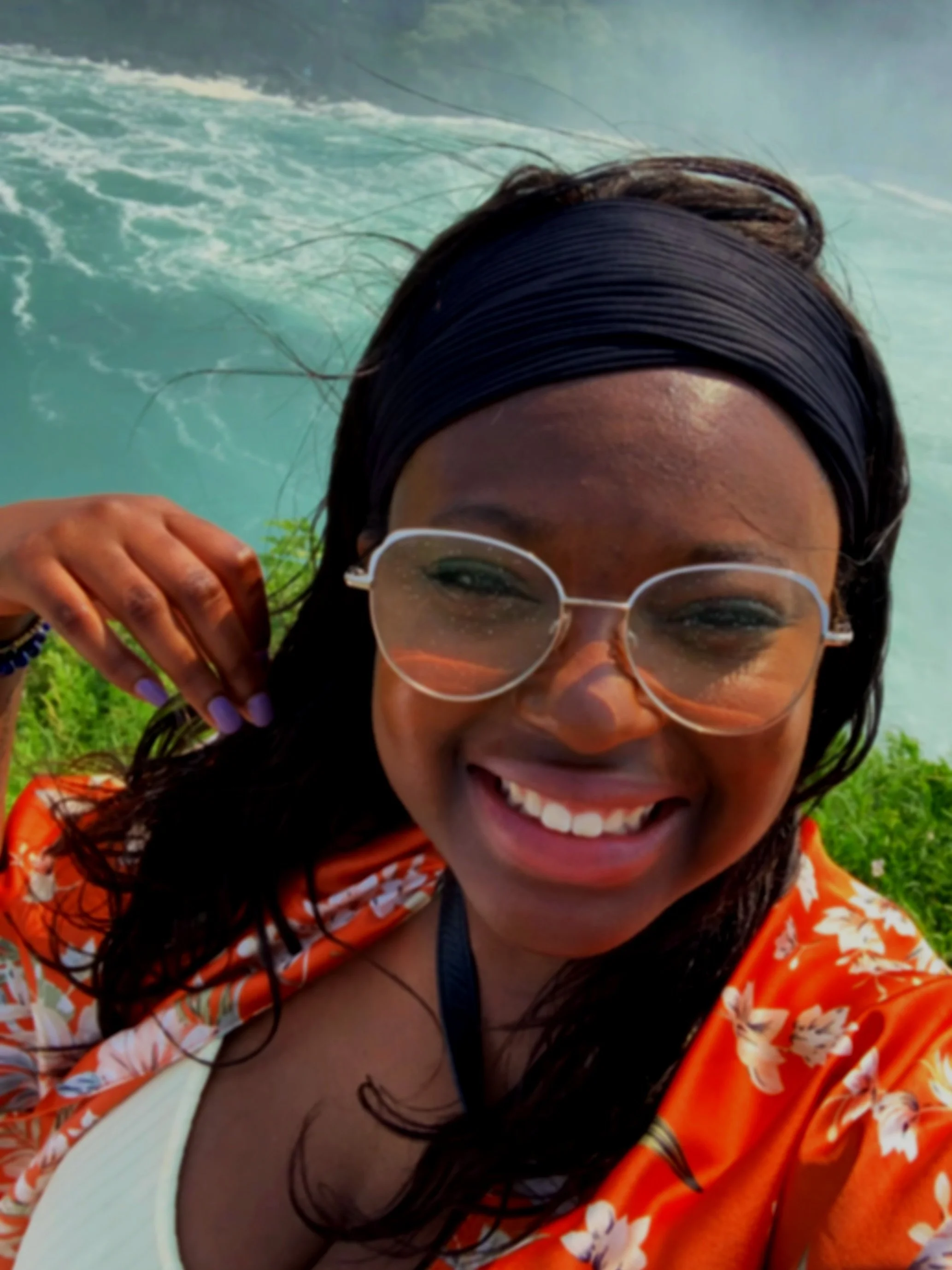 A smiling woman wearing glasses, a dark headband, and a bright floral orange shirt, taken outdoors near water.