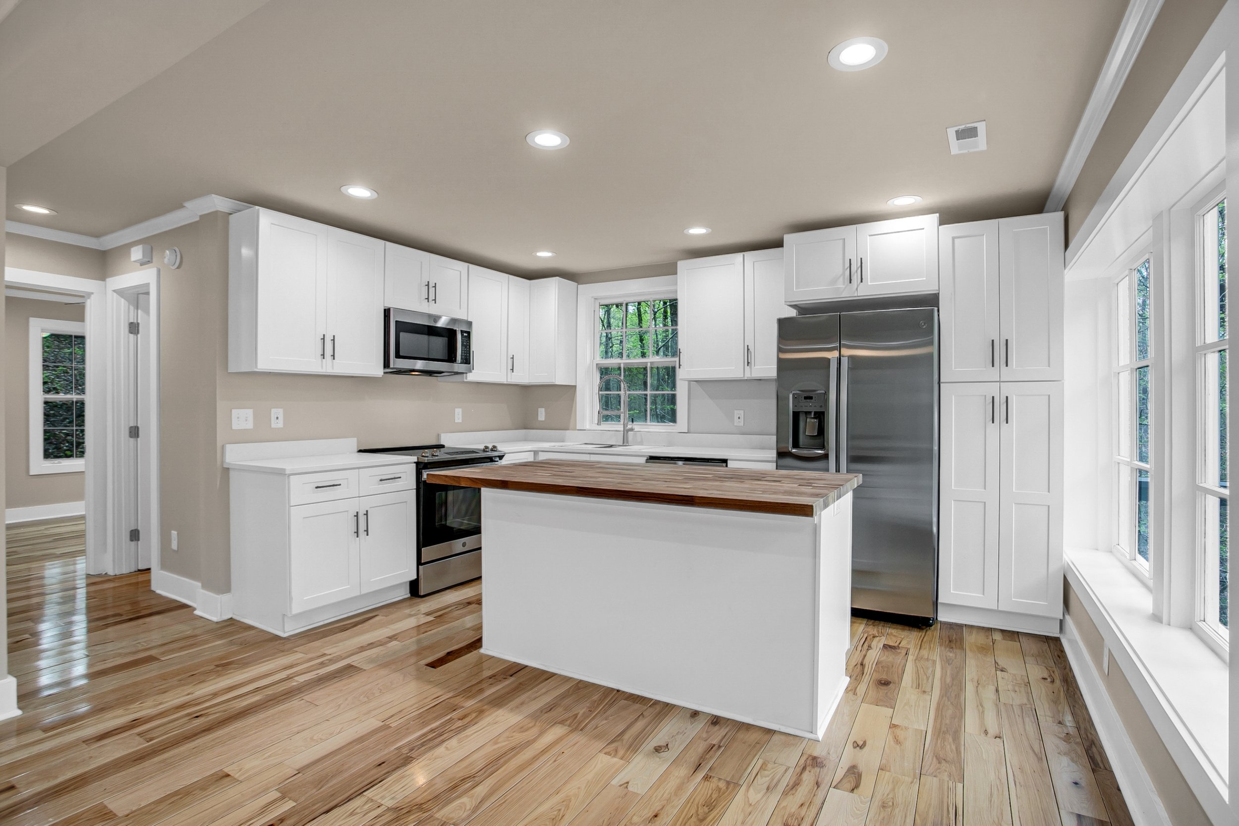 Modern white kitchen with stainless steel appliances, wooden flooring, and large windows overlooking greenery.