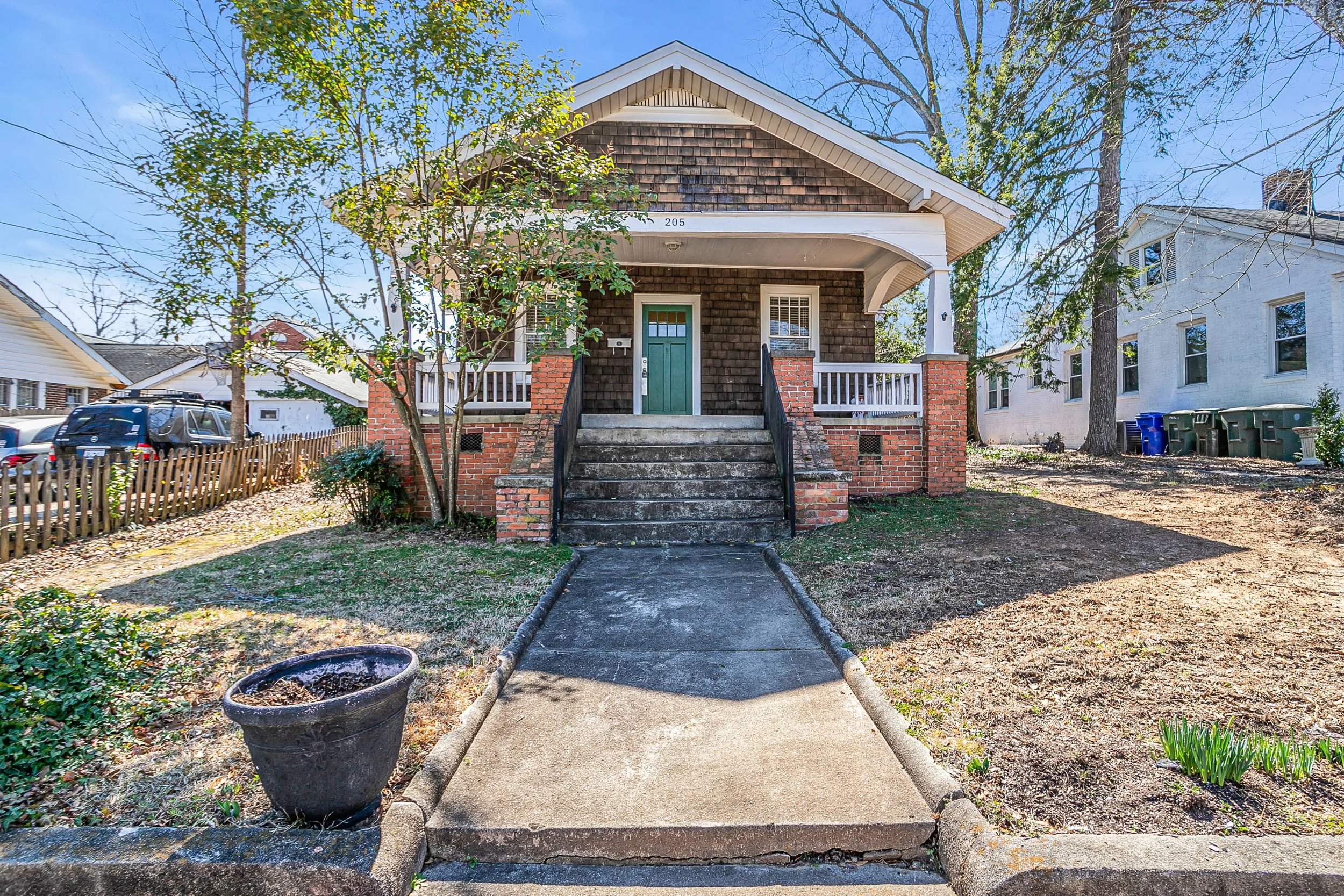 Front porch of a house with a green door, brick steps, and wooden shingles exterior. There are trees and neighboring houses, with a blue sky. A black cat is sitting on the steps.