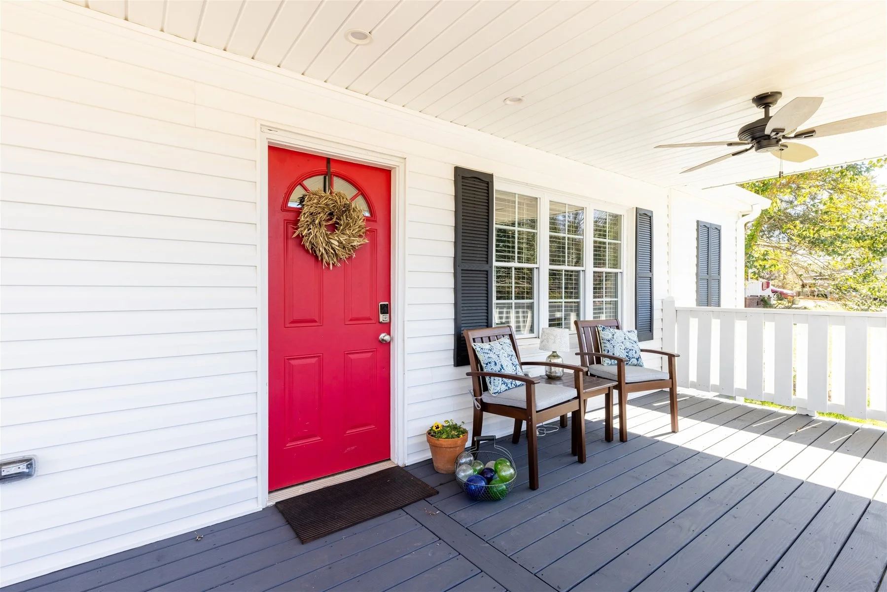 A porch with a red front door decorated with a straw wreath. There are two wooden chairs with cushions, a small table with a lamp, and a potted flower. The porch has white railing, black shutters, and a ceiling fan.