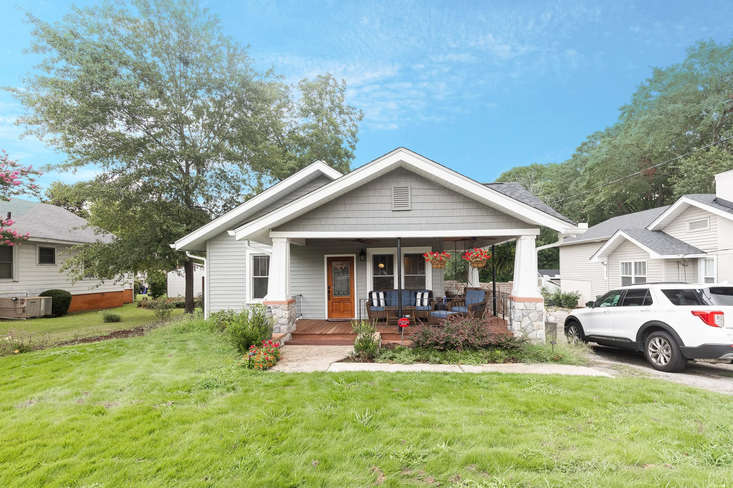 Front view of a white house with a porch, hanging flower baskets, and outdoor seating. Surrounded by green lawn and trees, with neighboring houses in the background and a clear blue sky.