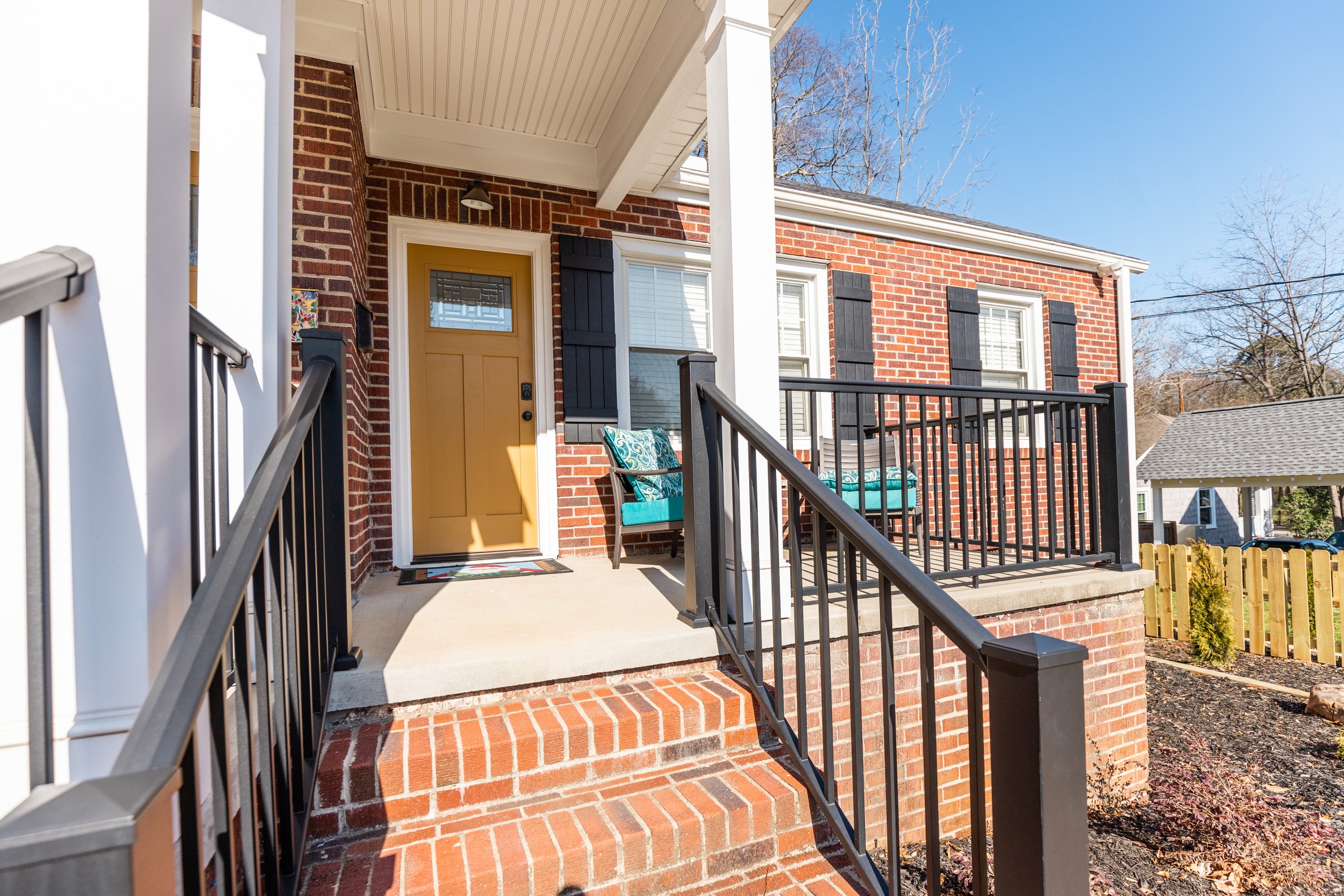 Front porch of a brick house with a yellow door, black shutters, and black railing stairs. Two chairs with turquoise cushions sit on the porch, and a colorful decorative item is on the wall. There are leafless trees and a clear blue sky in the background.