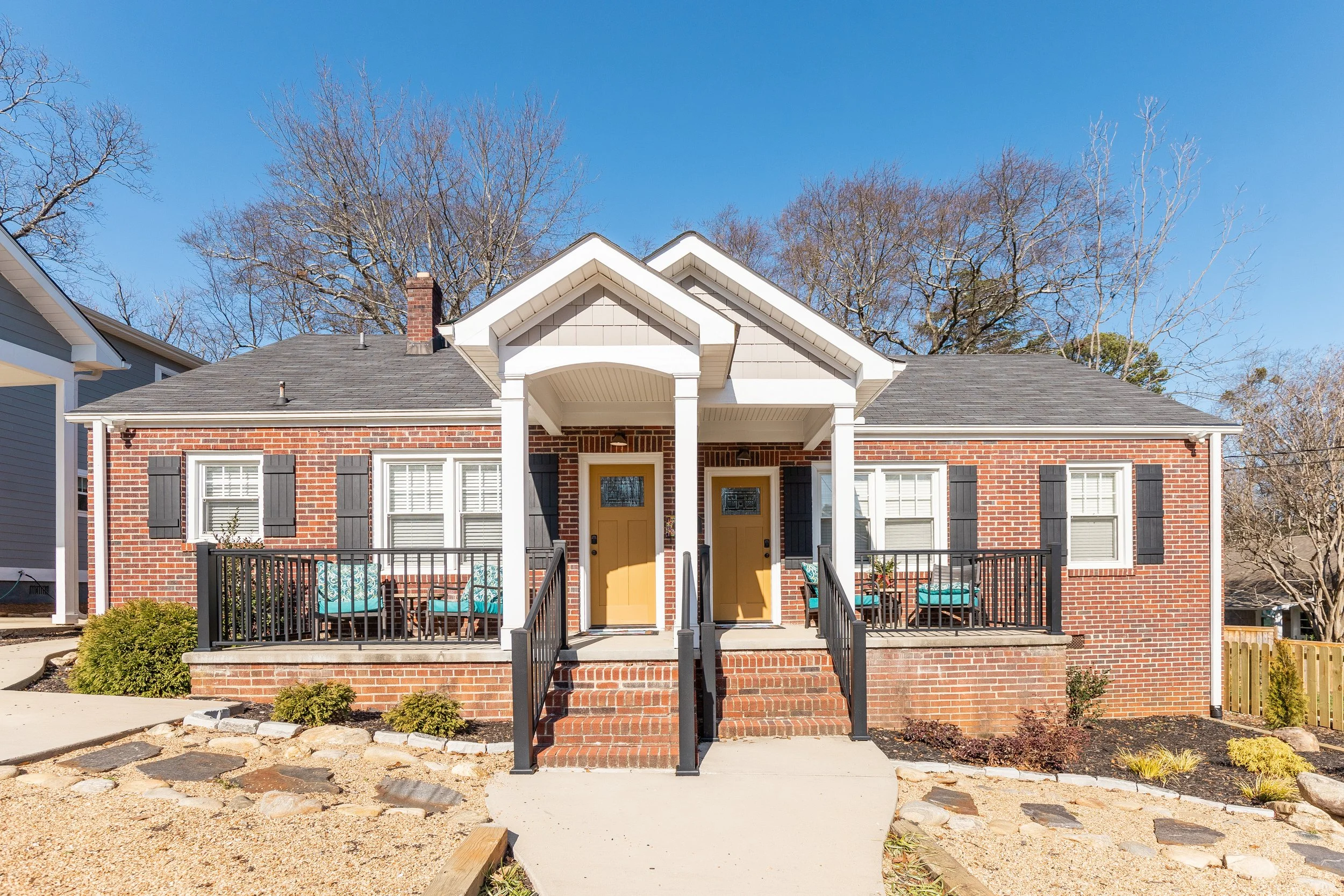 Front view of a brick house with a small porch, black railing, yellow front doors, and multiple windows with black shutters. There are two chairs on the porch, a small garden in front, and leafless trees in the background under a clear blue sky.