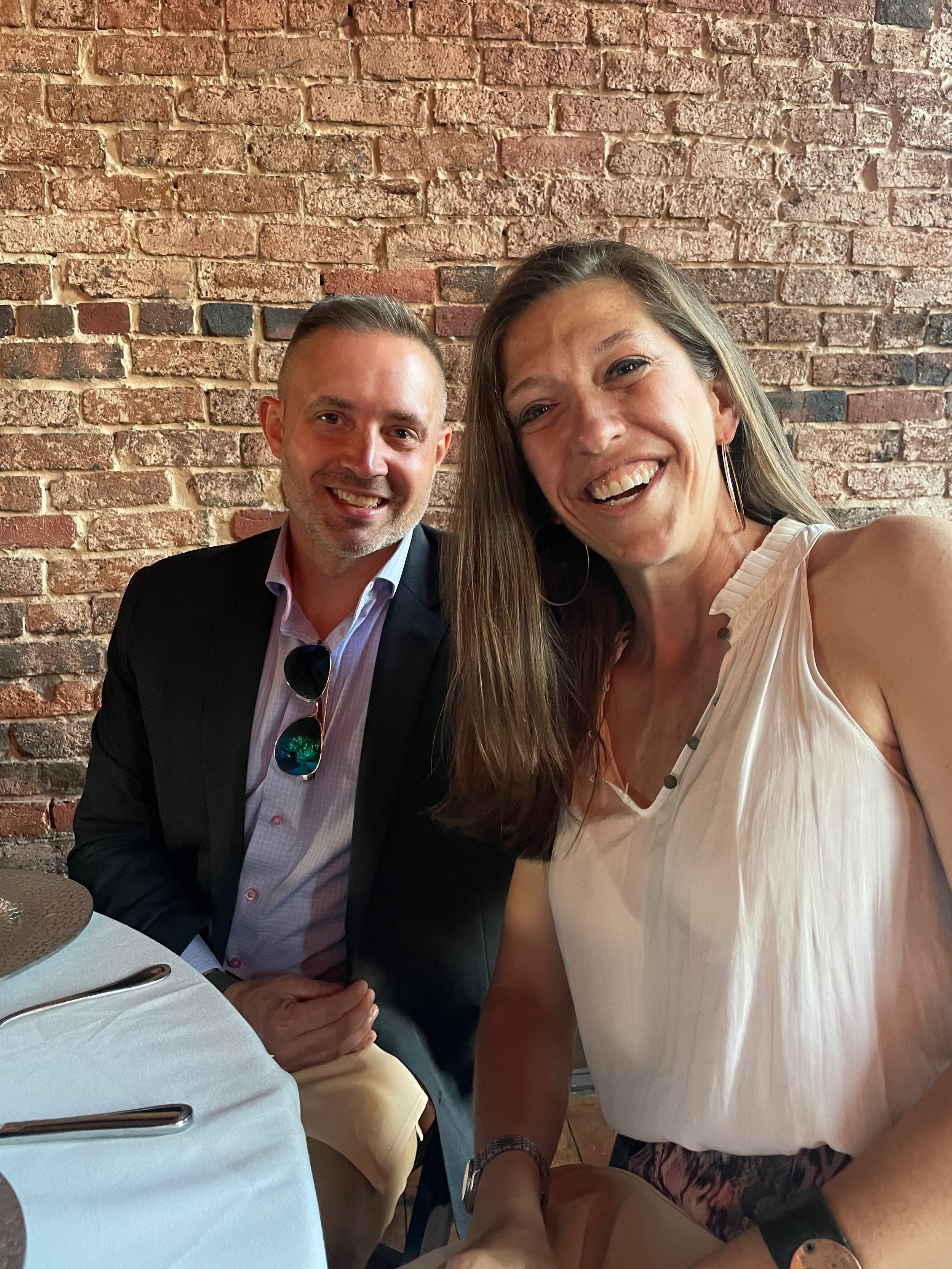 A woman and a man sitting at a restaurant table, smiling and taking a selfie with a brick wall in the background.
