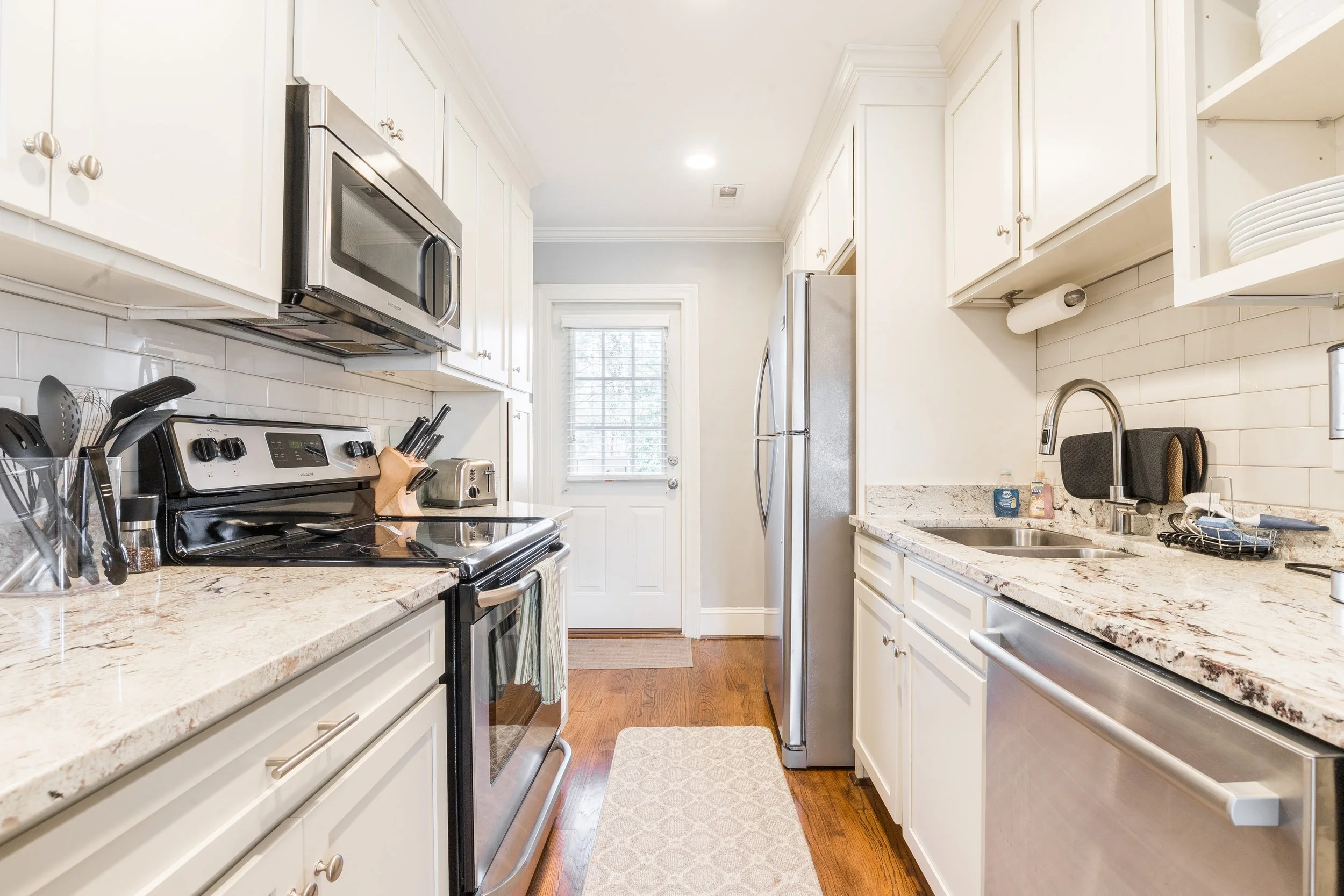 A kitchen with white cabinets, a granite countertop, and stainless steel appliances including a microwave, oven, and refrigerator. There is a window at the back door with blinds, a sink with a faucet, and various kitchen items on the counter.