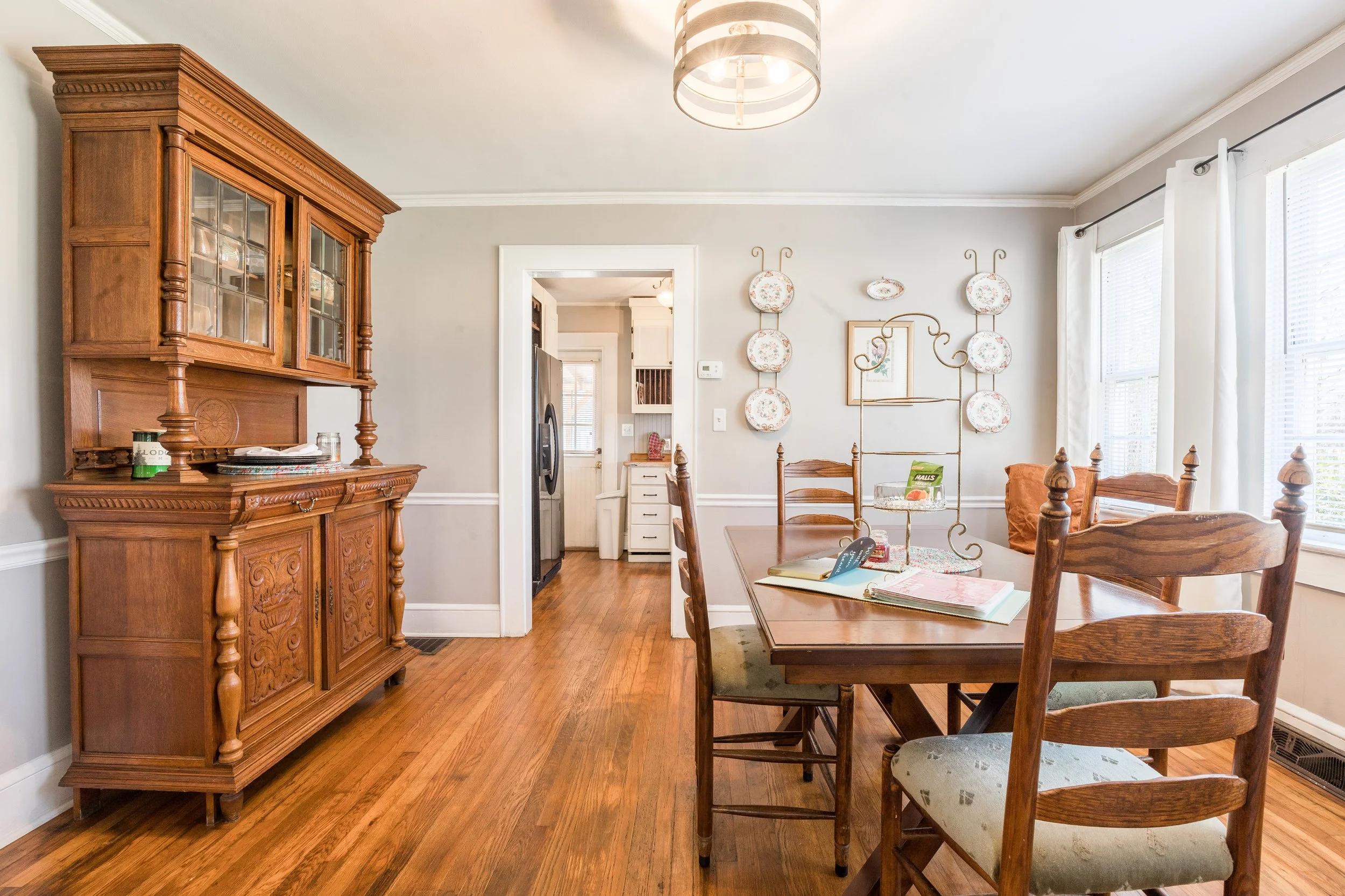 A cozy dining room with a wooden table and six matching chairs. A large antique wooden china cabinet is on the left, and a wall decorated with floral plates is on the right. A doorway in the middle leads to a kitchen area, with a washer and dryer visible. Large windows with white curtains allow natural light into the space. The room has hardwood flooring and neutral-colored walls.