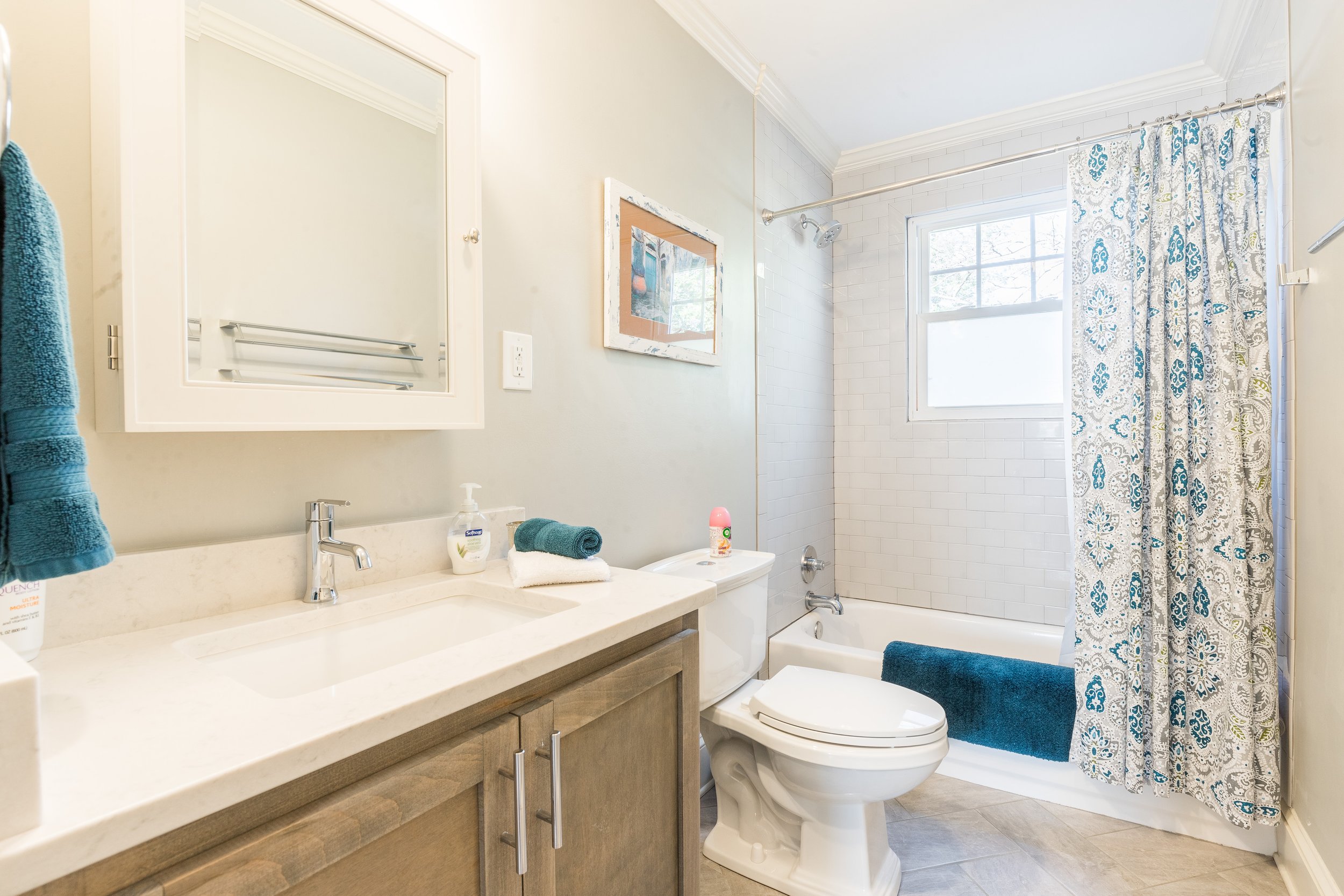 A bathroom with a beige vanity, a white countertop, a mirror, a hand soap dispenser, and towels. There is a toilet and a bathtub with a shower curtain featuring blue and gray patterns. A window provides natural light.