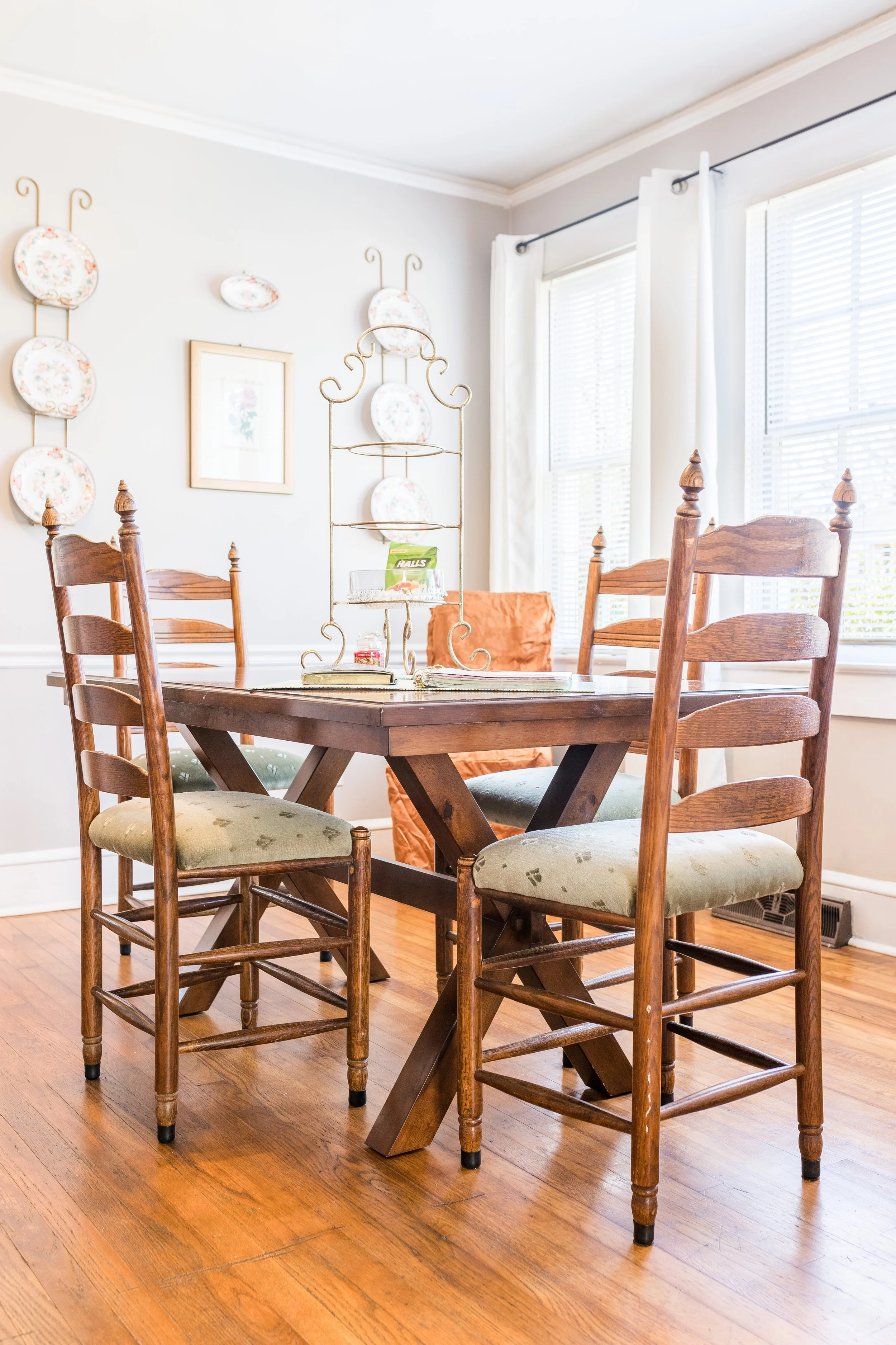 A cozy dining room featuring a wooden dining table surrounded by four wooden chairs with upholstered seats. The room has hardwood flooring, a white wall decorated with floral plates and a framed picture, and large windows with white curtains allowing natural light to enter.