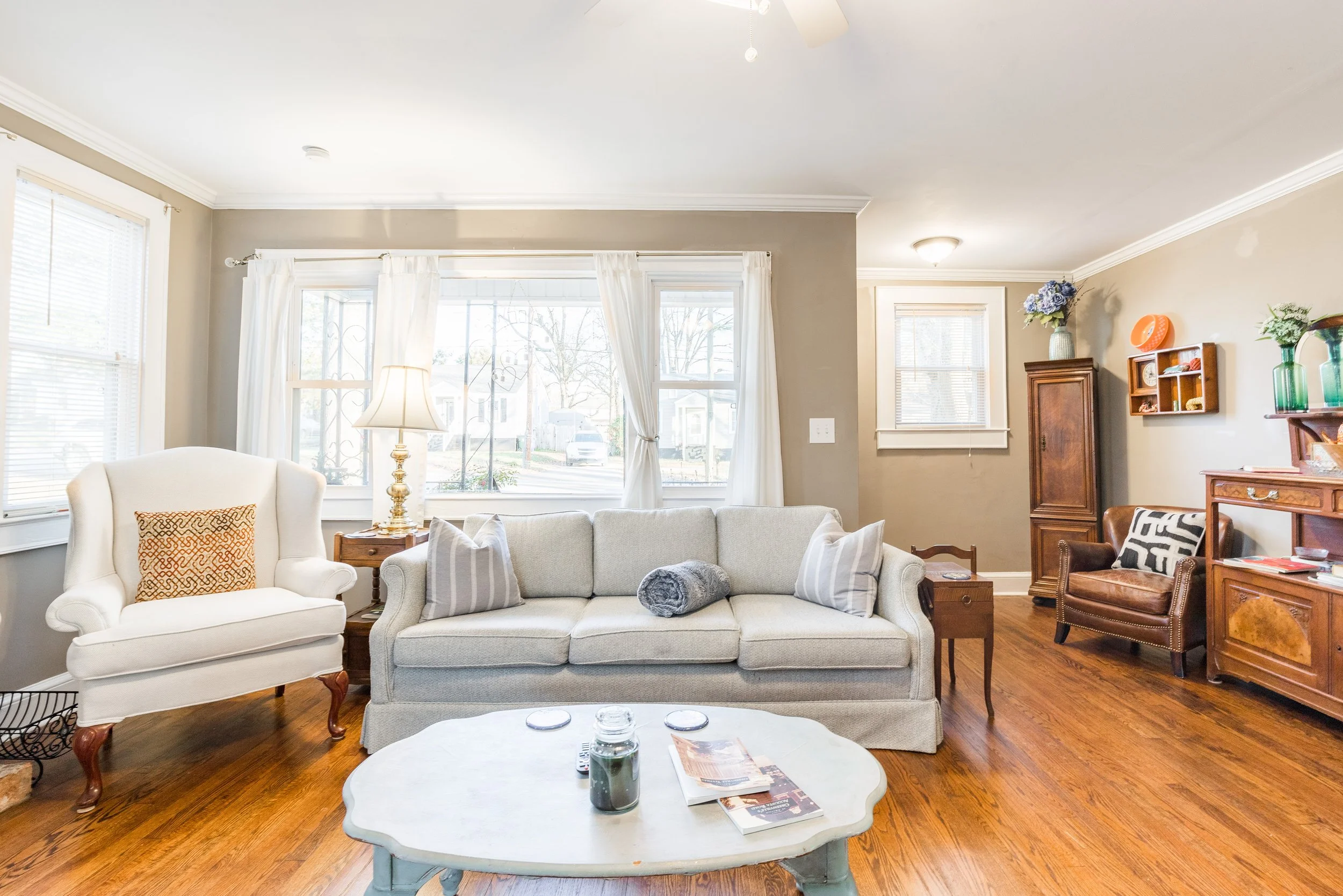Living room with large windows, beige walls, white curtains, cream-colored sofa, armchair with decorative pillow, wooden side table, lamps, and wooden furniture including a chest of drawers and a small wall shelf with vases and decorative items. Hardwood floors.