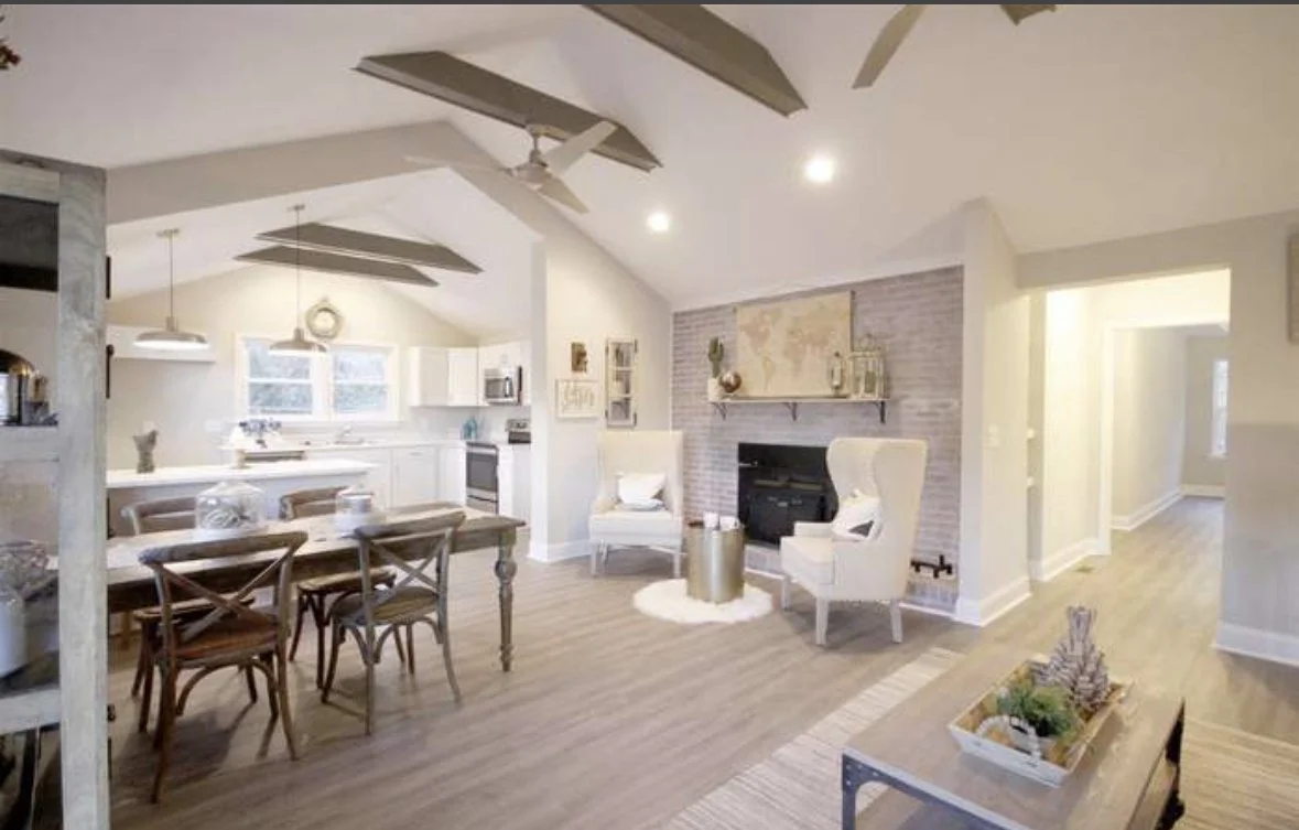 Open-concept living and dining area with high vaulted ceiling, hardwood floors, and neutral decor. The living room features two white armchairs, a black fireplace with a brick accent wall, a world map, and shelves with decorations. The dining area has a wooden table with six chairs, overhead pendant lights, and positioned near a kitchen with white cabinets and appliances.