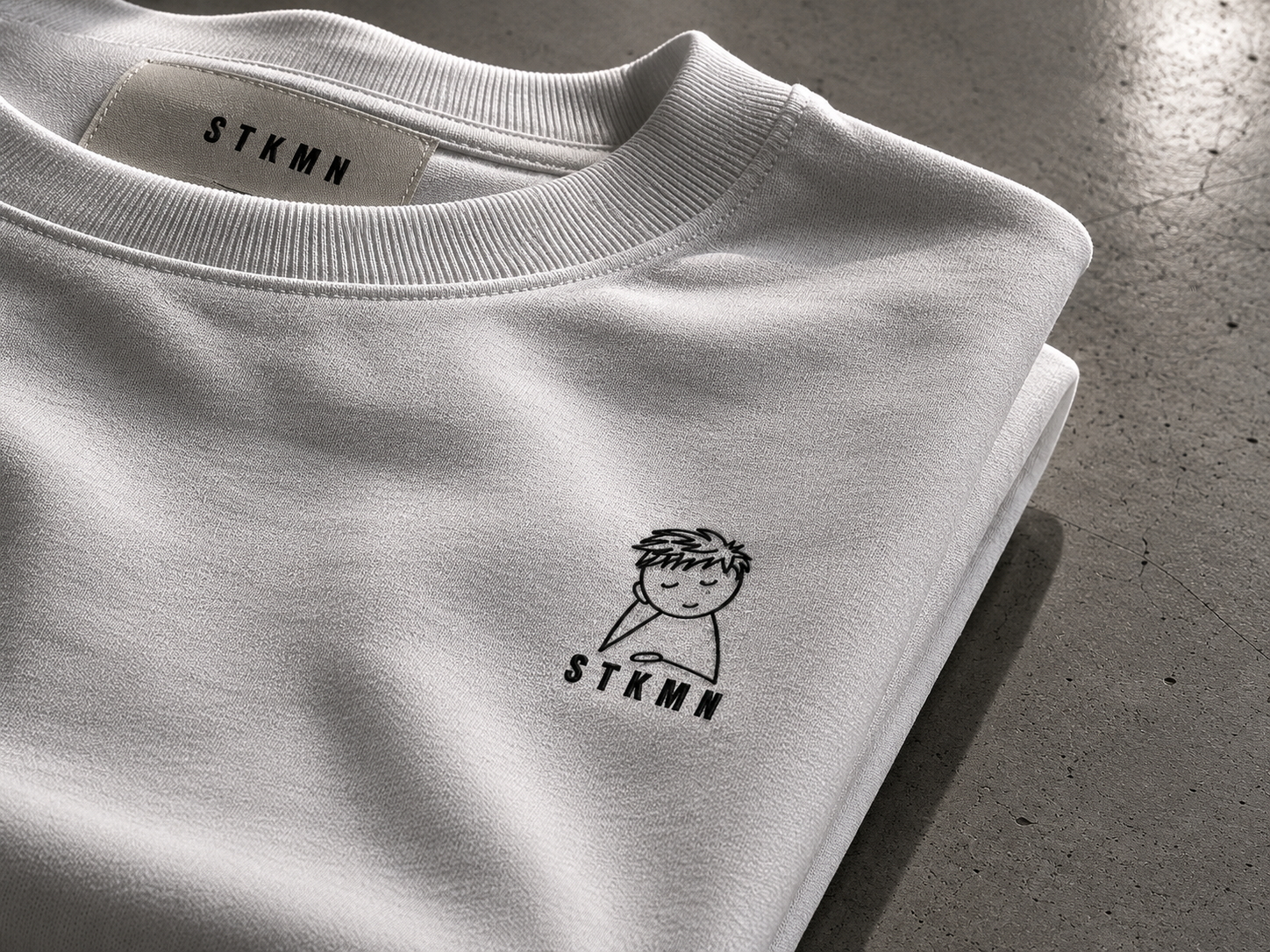 A white sweatshirt with a small cartoon character of a smiling boy with spiky hair and closed eyes, holding a pencil, printed on the bottom left side. The collar of the sweatshirt is visible and the background is a concrete surface.