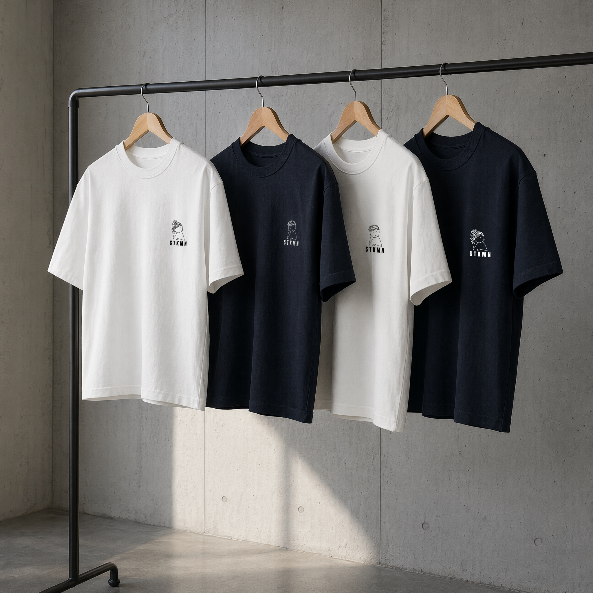 A clothing rack displays four T-shirts, two white and two black, each on a wooden hanger, against a concrete wall background.