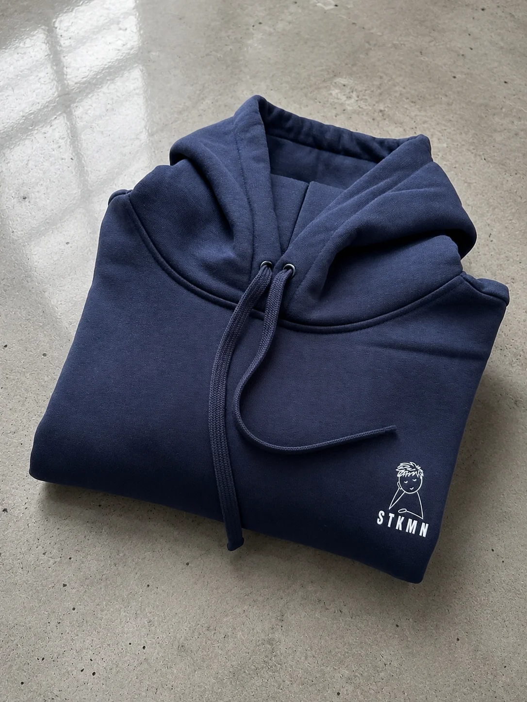 A folded navy blue hoodie with a white logo of a smiling face and the letters 'STKMN' on the sleeve, placed on a concrete surface.