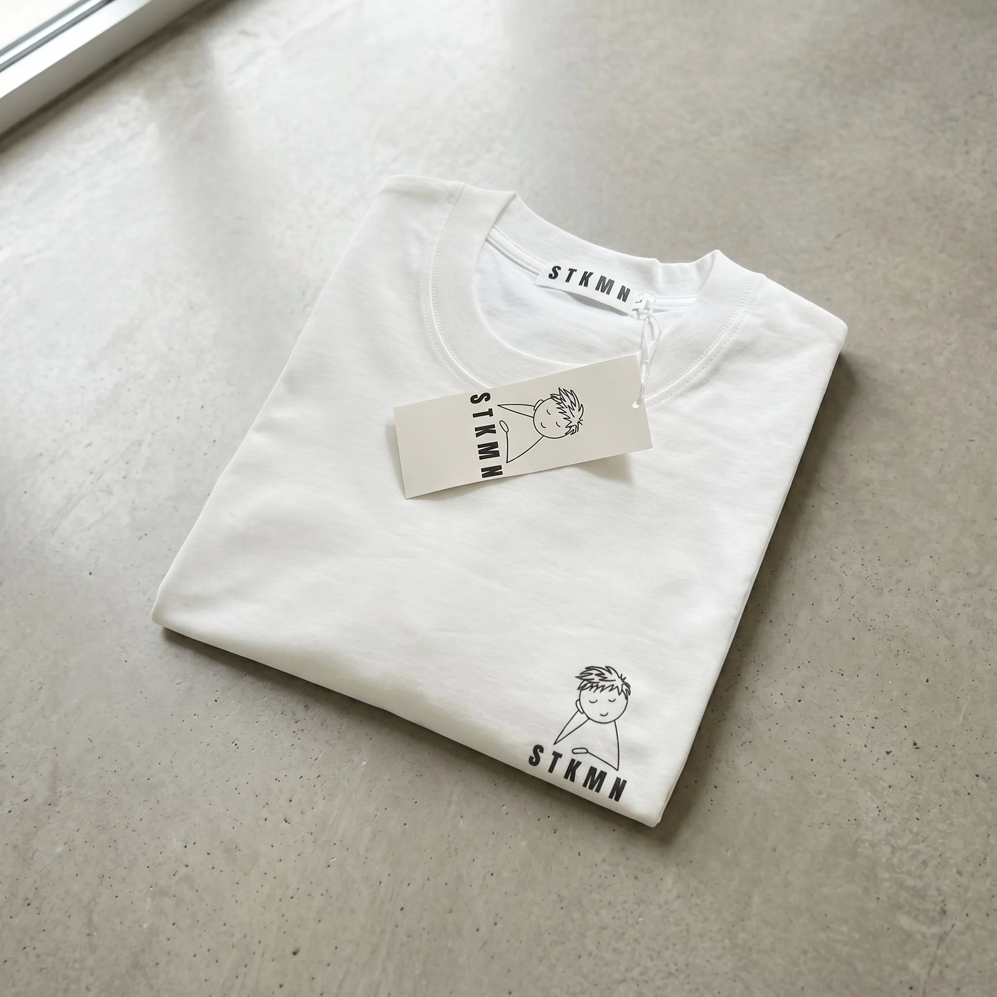 A folded white t-shirt with a simple black graphic and text, placed on a light-colored speckled surface, near a window.