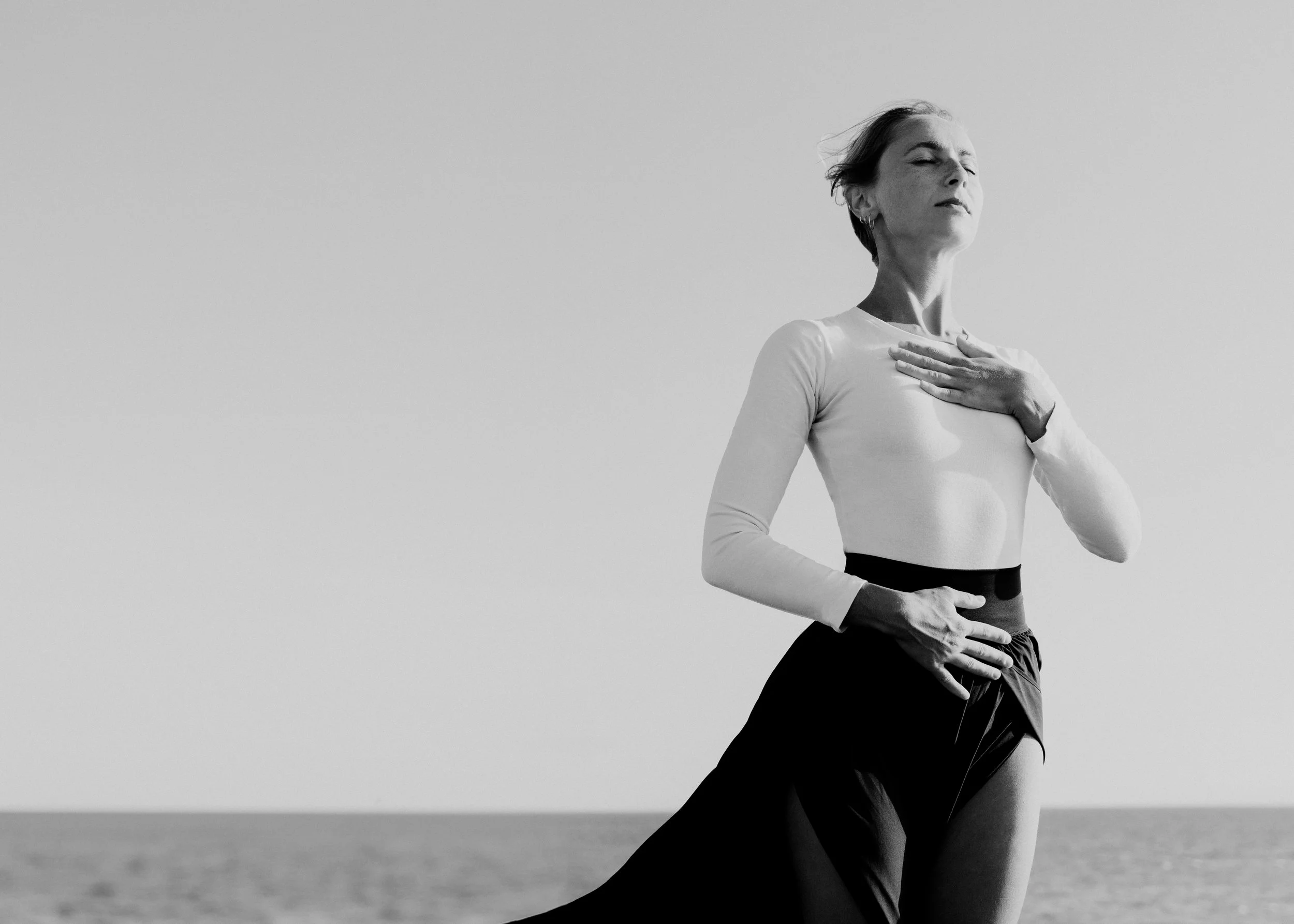 Woman with her hands in her heart and belly, breathing freely in nature with the icean behind her.