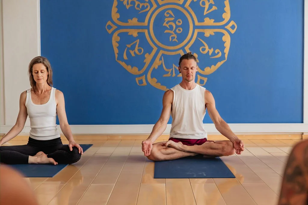 a woman and a man practice meditation in crossed-legged posture in front of a purple wall.