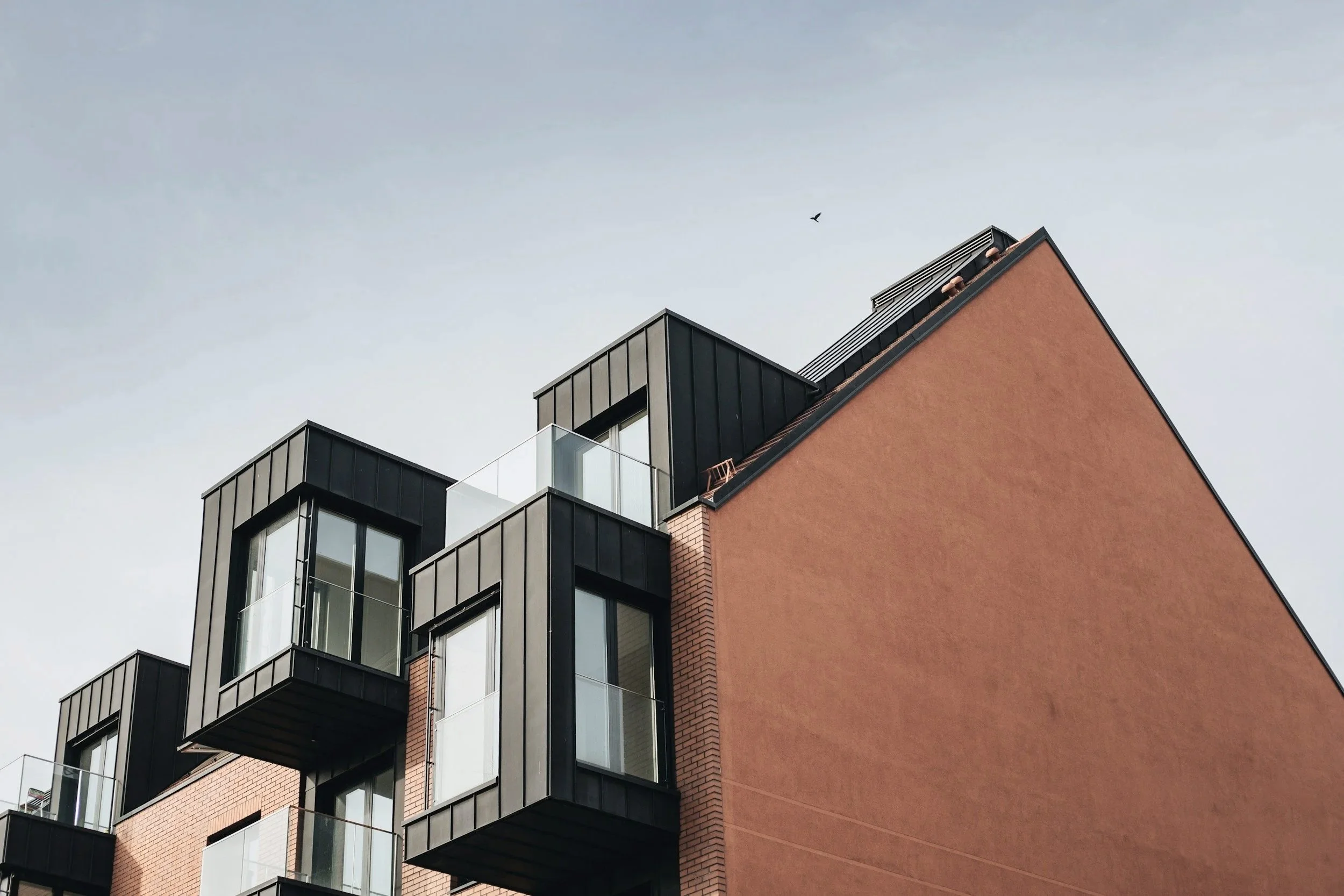 Photo d'un bâtiment moderne avec plusieurs balcons en verre et en métal, et une façade en briques roses, vue en contre-plongée contre un ciel clair.