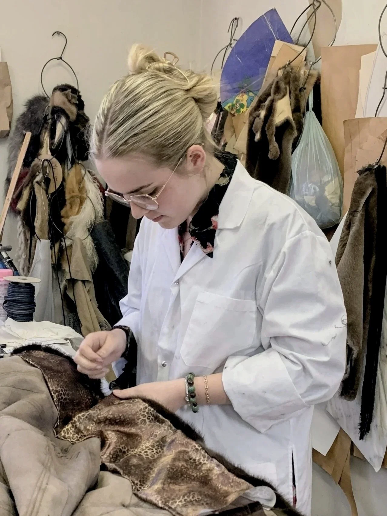 A woman in a white lab coat and glasses working on a plush animal with leopard print fabric in a workshop filled with fur, fabric, and craft supplies.