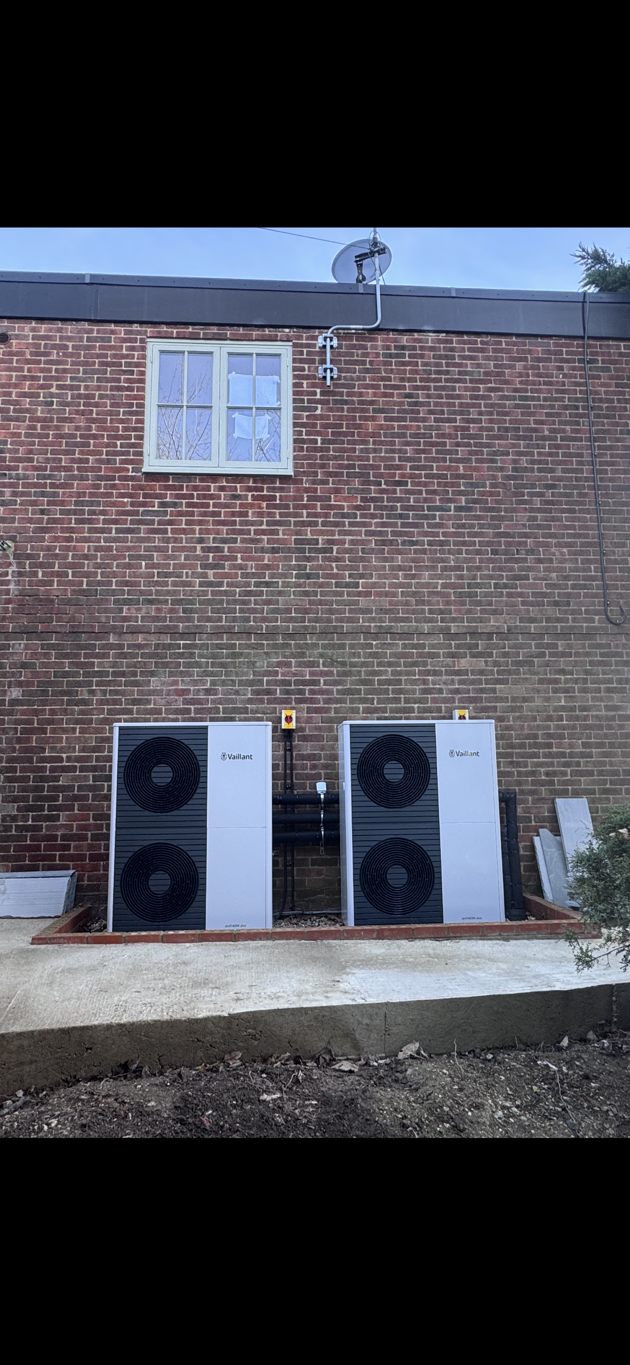 Two outdoor heat pump units labeled Vaillant arotherm plus installed on a brick wall ground in a backyard, with a window and satellite dish above.